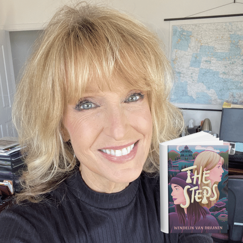 A woman with blonde hair and a black shirt smiling while holding a book titled 'The Steps' by Wendelin Van Draanen. She is in an office with a world map on the wall, a computer, and stacks of papers or files.