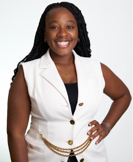 Smiling woman with braided hair wearing a sleeveless white blazer, black top, gold necklace, standing with one hand on her hip.