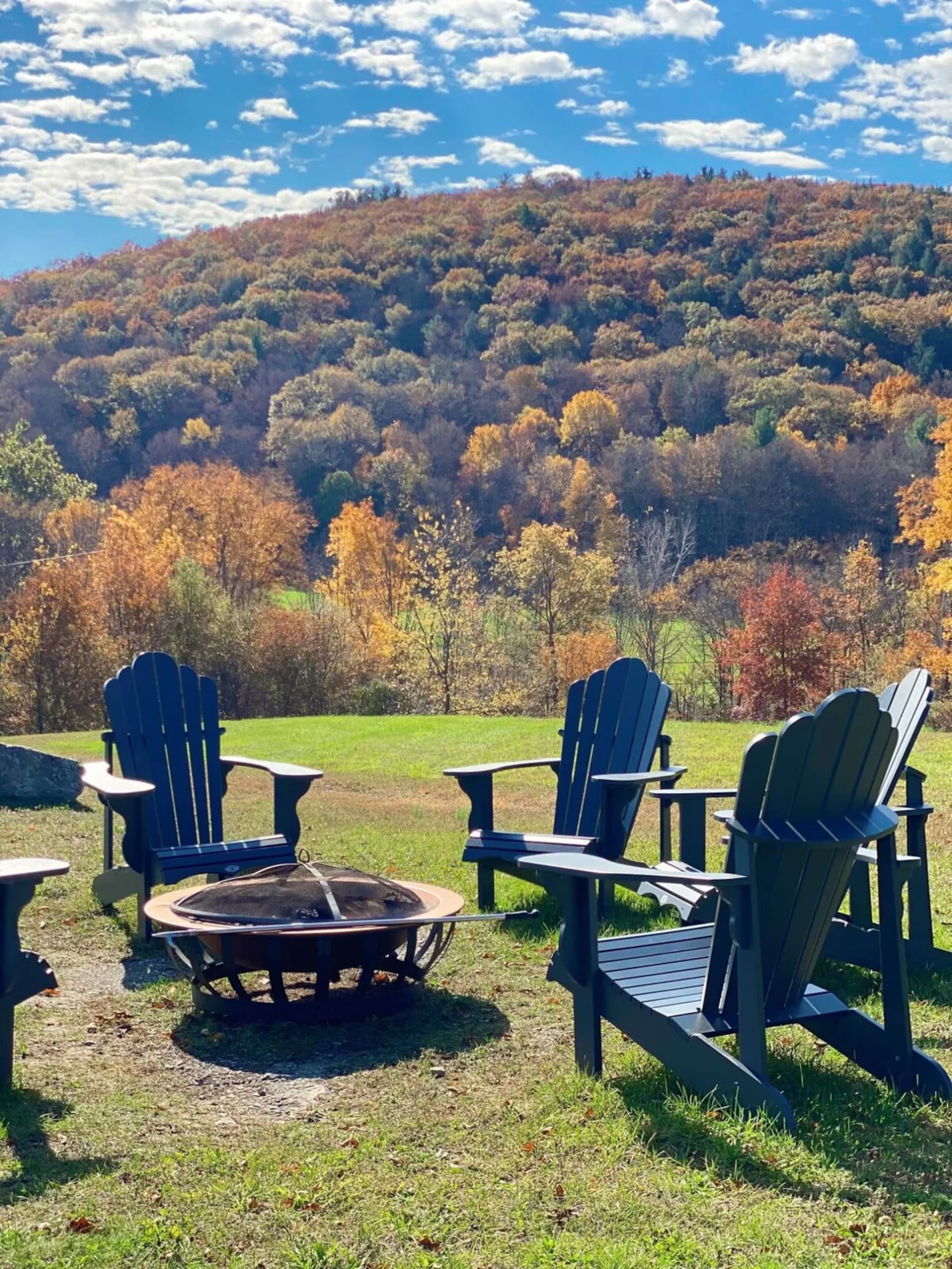 A scenic outdoor view featuring a grassy area with several blue Adirondack chairs arranged around a fire pit. Behind the chairs, there are colorful trees with fall foliage and a mountain with trees in the distance. The sky above is partly cloudy with patches of blue.