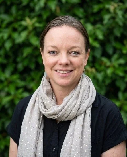A woman with light brown hair pulled back, wearing a black shirt and a light gray scarf with white polka dots, smiling in front of green foliage.