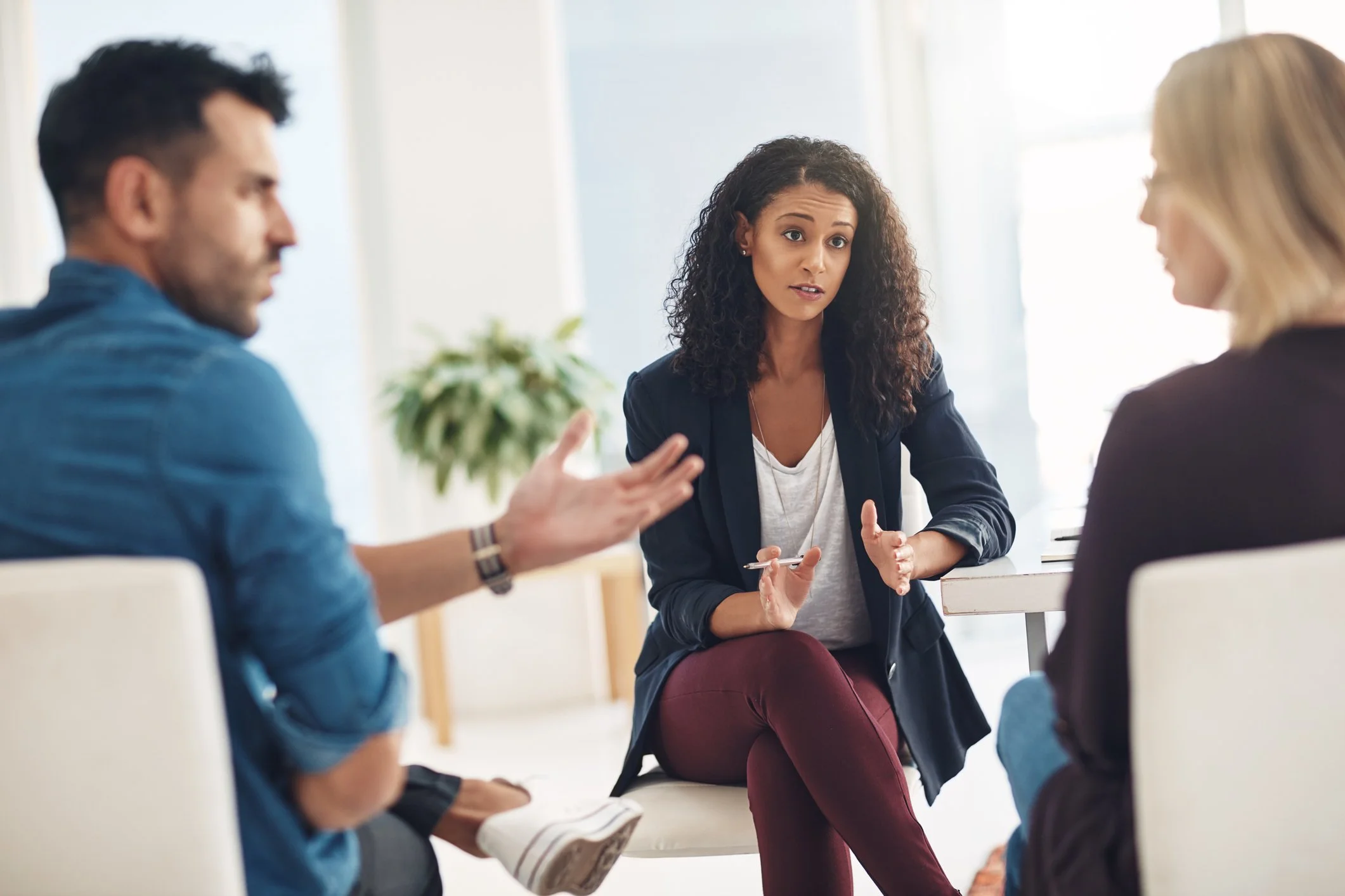 A woman in a dark blazer and maroon pants speaks during a group discussion with two other women and a man in a bright room with large windows and a potted plant in the background.
