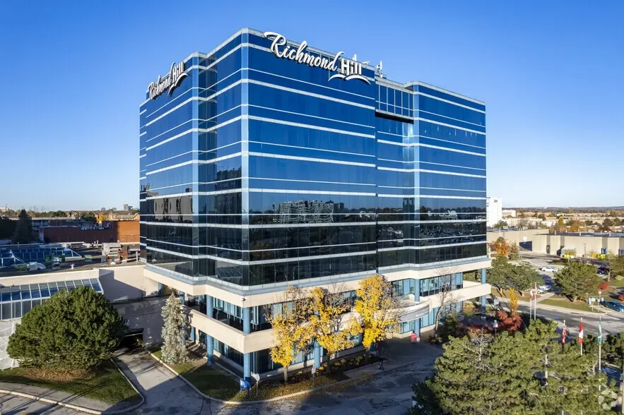 A tall modern glass office building with the sign Richmond Hill on top, surrounded by trees and parking lots under a clear blue sky.