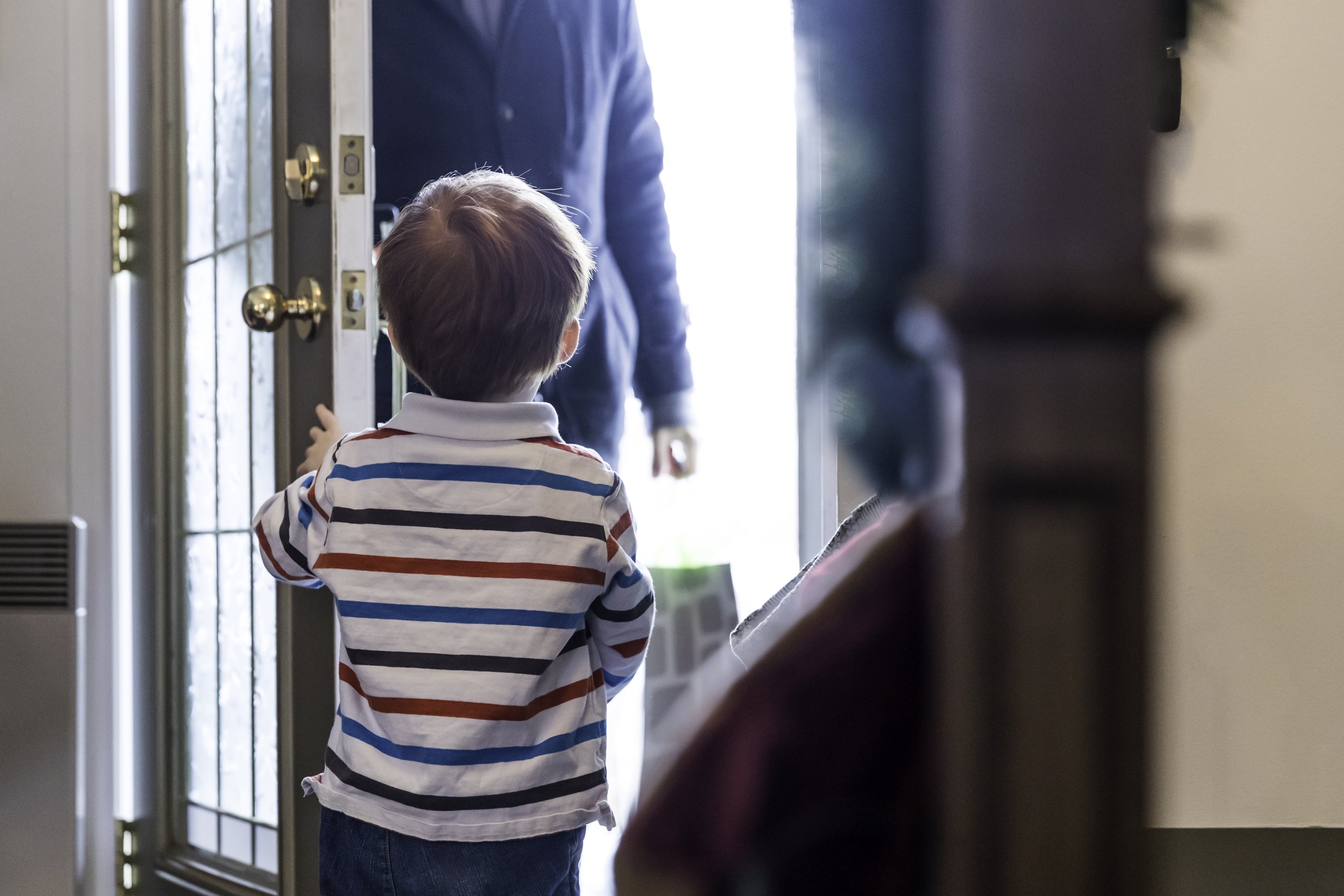 A young boy with striped shirt reaching for the door to greet an adult, perhaps his father or mother, standing outside the door. A stock photo representative of themes of parenting-time and difficulties of divorce.