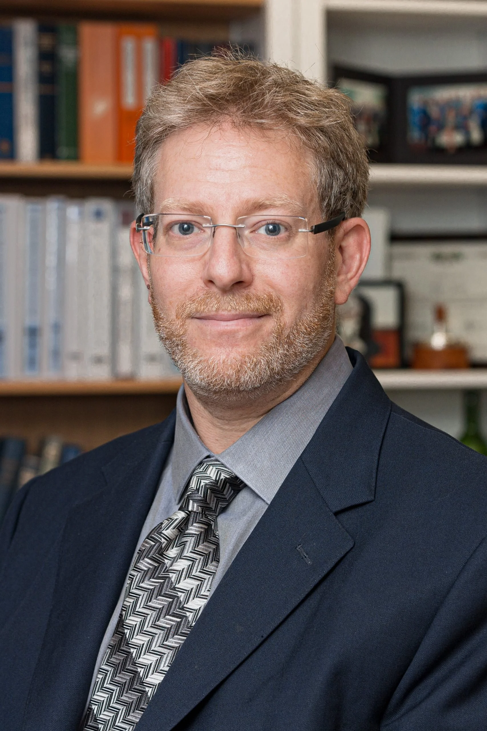 Professional headshot of Avram Joseph, a senior family lawyer, in formal attire, suit, tie and glasses, with shelves of books in the background behind him.