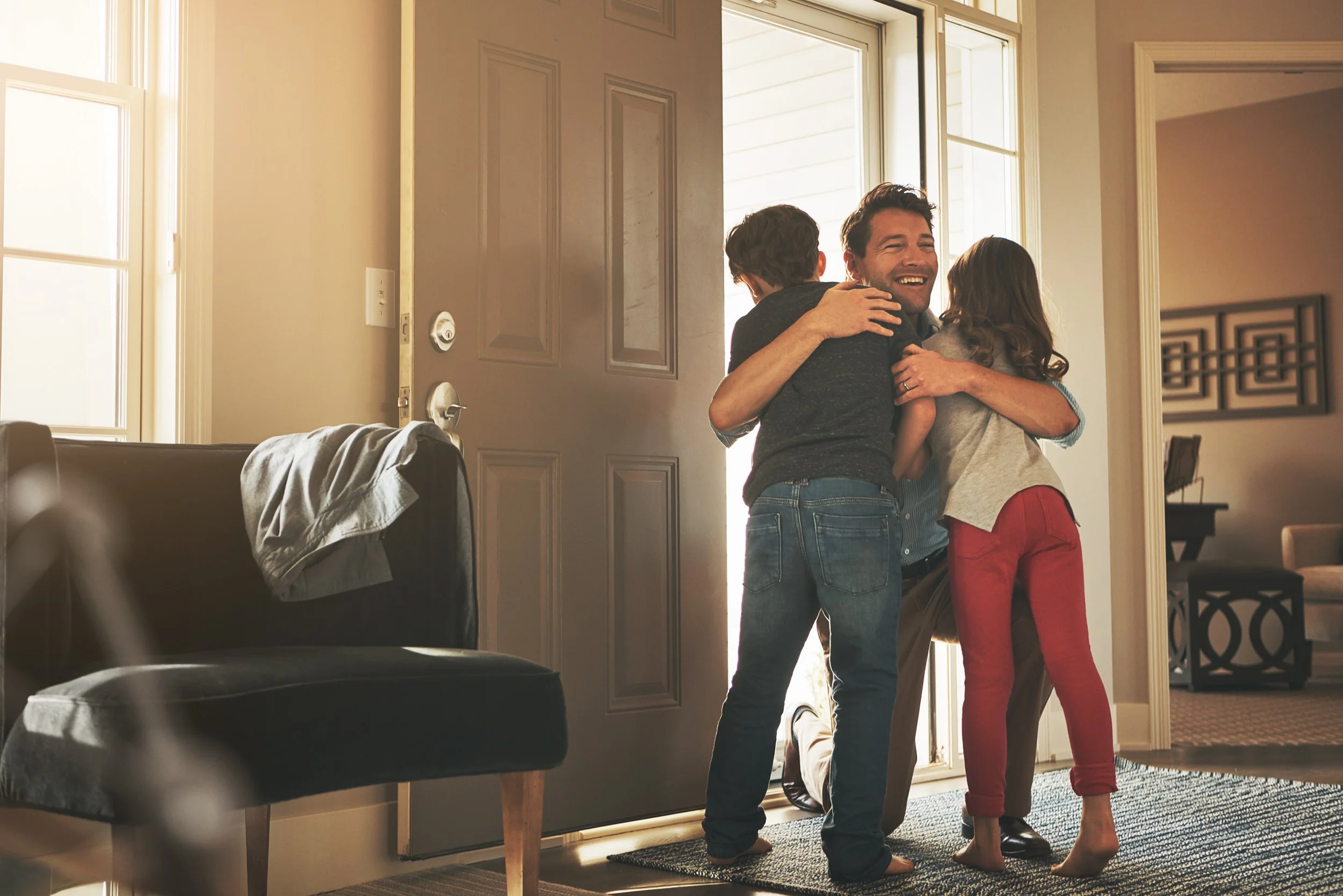Kids welcome their dad home inside in front of the open front door in a living room or hallway type space. A stock photo.
