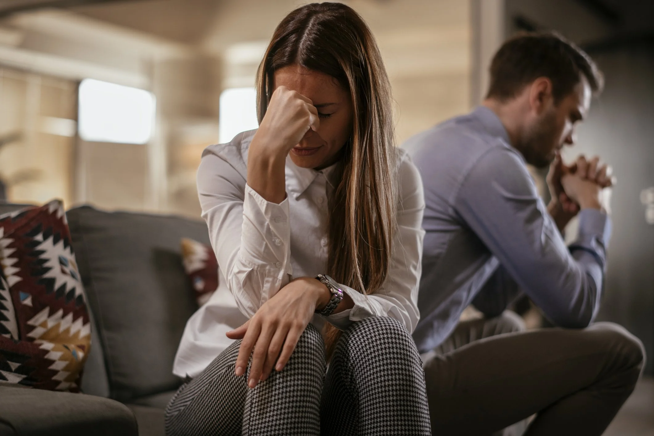 A woman with long hair is sitting on a couch, upset, holding her forehead. A man with short brown hair is sitting next to her, also upset, with his hands clasped. A stock photo on theme of marriage falling apart into separation and divorce.