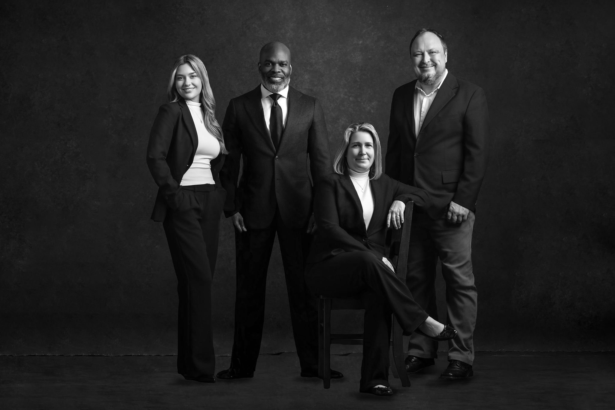 Black and white photo of four professionally dressed people, three men and one woman, posing against a dark textured background. The woman is seated on a chair and the others are standing around her. They are smiling and looking at the camera.