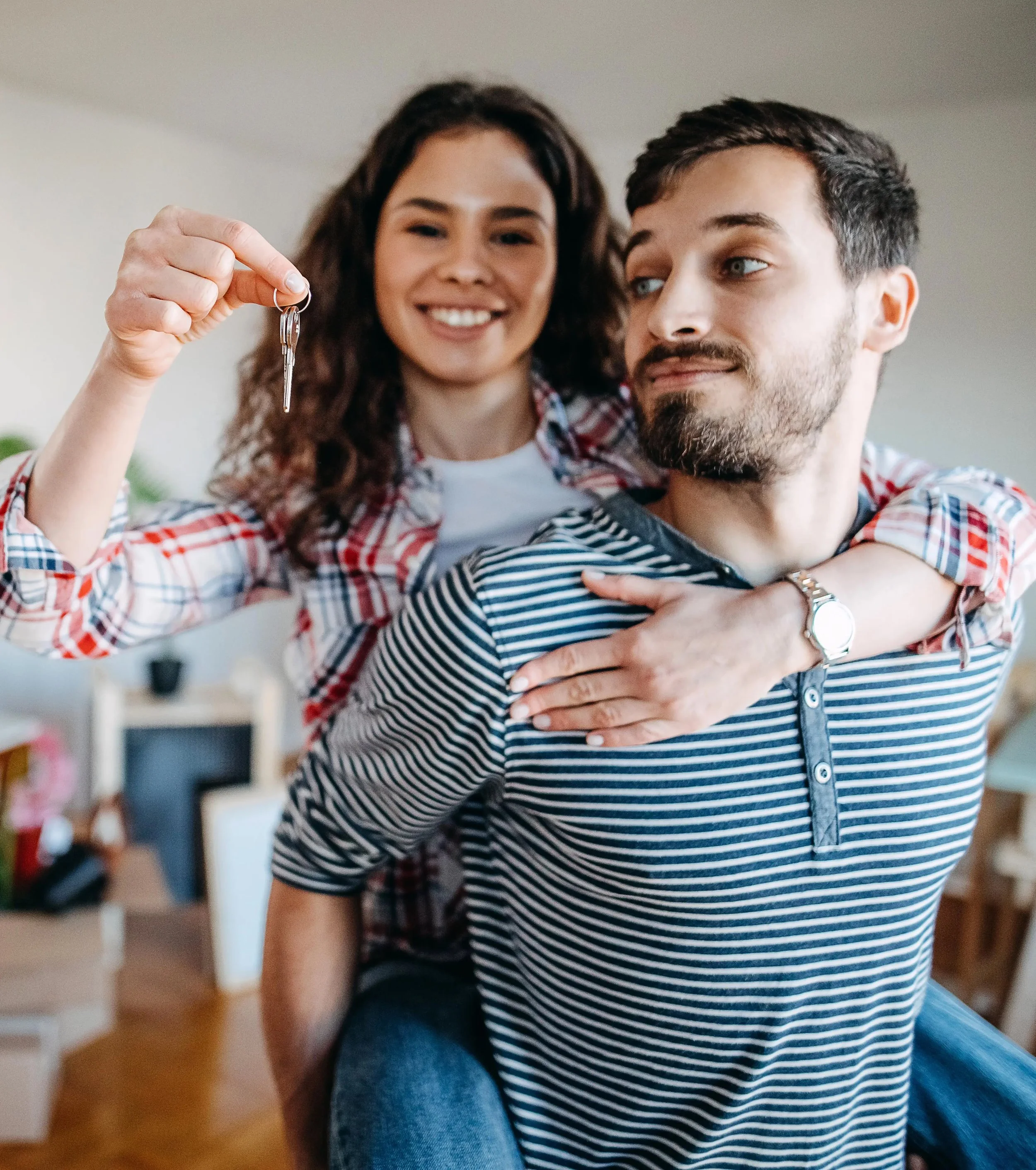 A smiling woman riding on a man's back in a cozy living room, holding a keys, perhaps to her new home or another real estate property. A stock image.