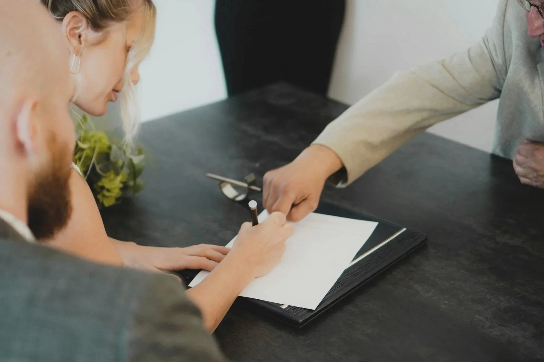 Business meeting with a man pointing at a signed document on the table while a woman signs it.