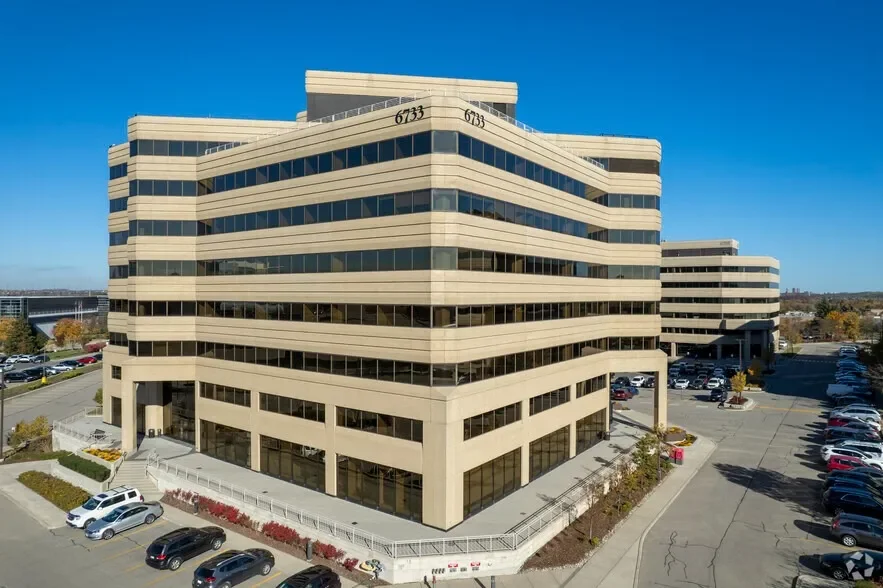 A large multi-story office building with beige exterior and horizontal dark-tinted windows, numbered 6733, situated in a parking lot with several cars, under a clear blue sky.