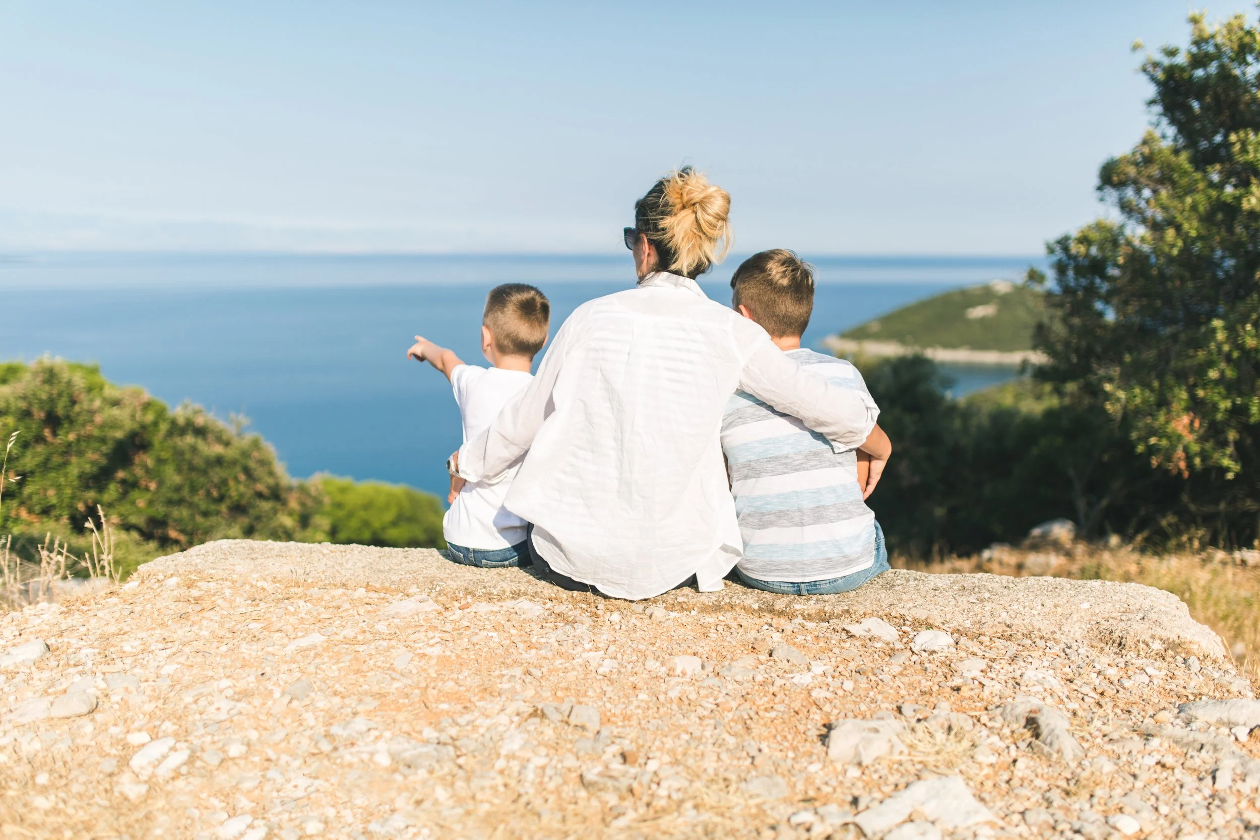 A mother with two young children sitting on a rock overlooking a body of water and green hills, with one boy pointing towards the distance. A stock photo in the context of a page on separation, parenting time or divorce.
