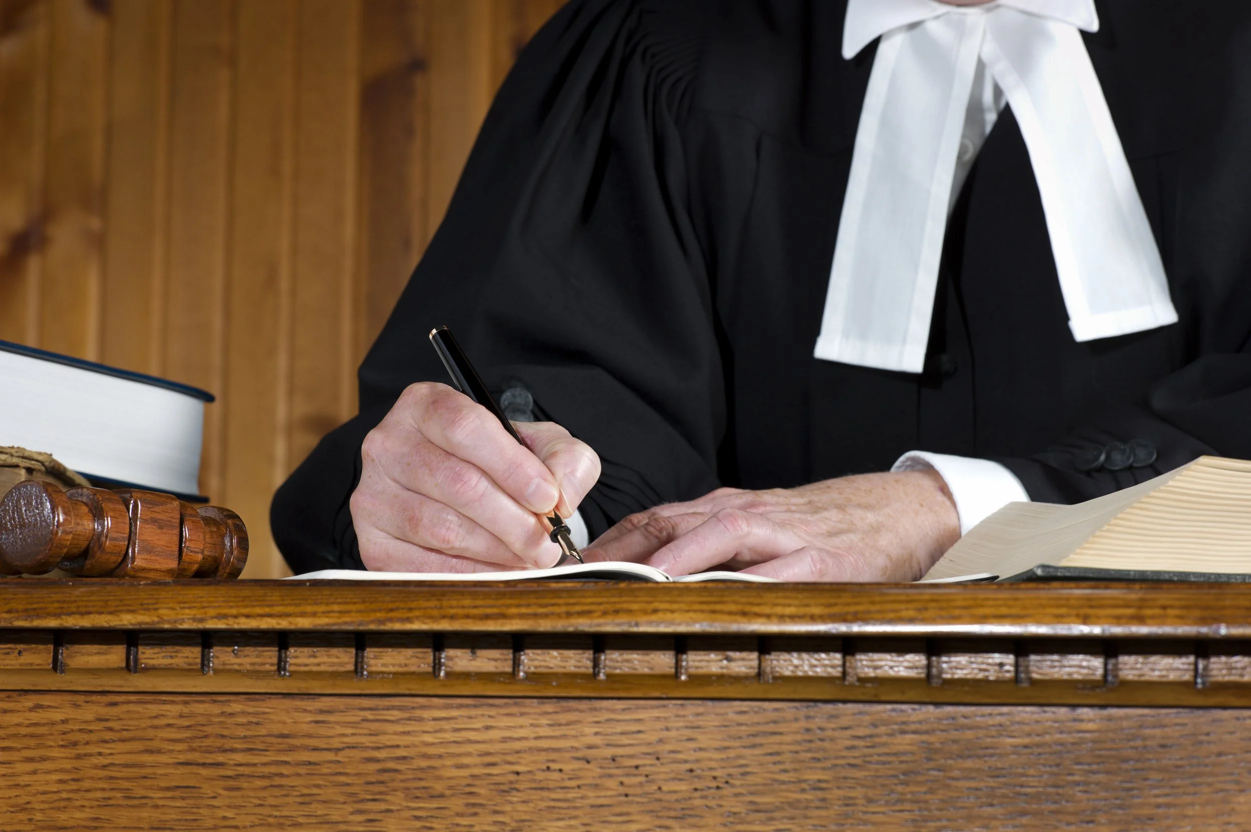 A judge in a court of law, wearing a black robe with a white collar and a white tie, writing in a legal document with a pen at a wooden desk with legal books and a gavel.