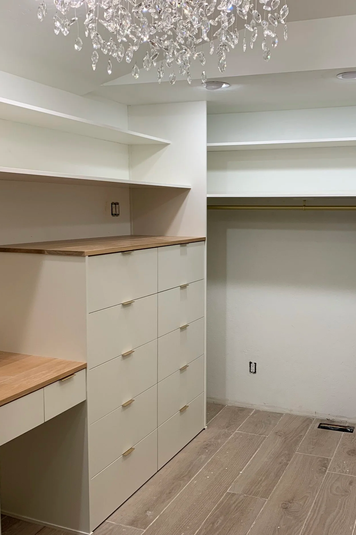 An empty walk-in closet with white built-in drawers, open shelves, and a hanging rod. A chandelier hangs from the ceiling, and the floor is tiled with large, wood-look tiles.