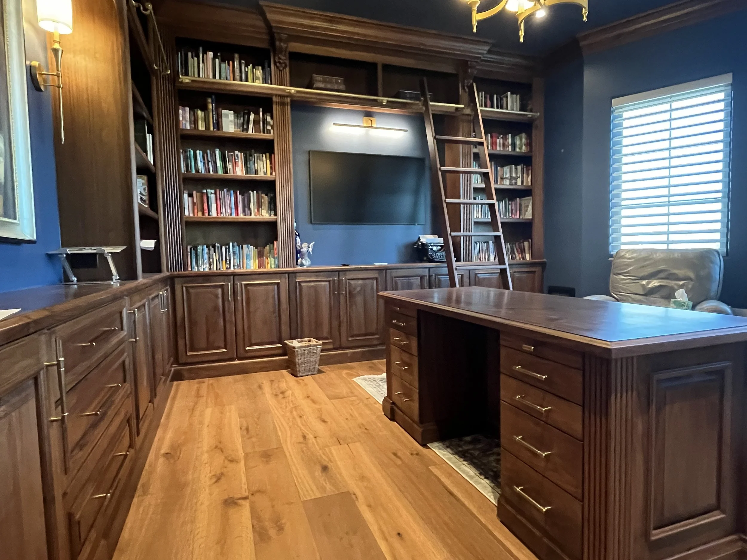 A home office or library with dark wooden built-in bookshelves, ladder, and furniture. There is a large flat-screen TV on the central wall, a window with blinds, a leather chair, and wooden flooring.