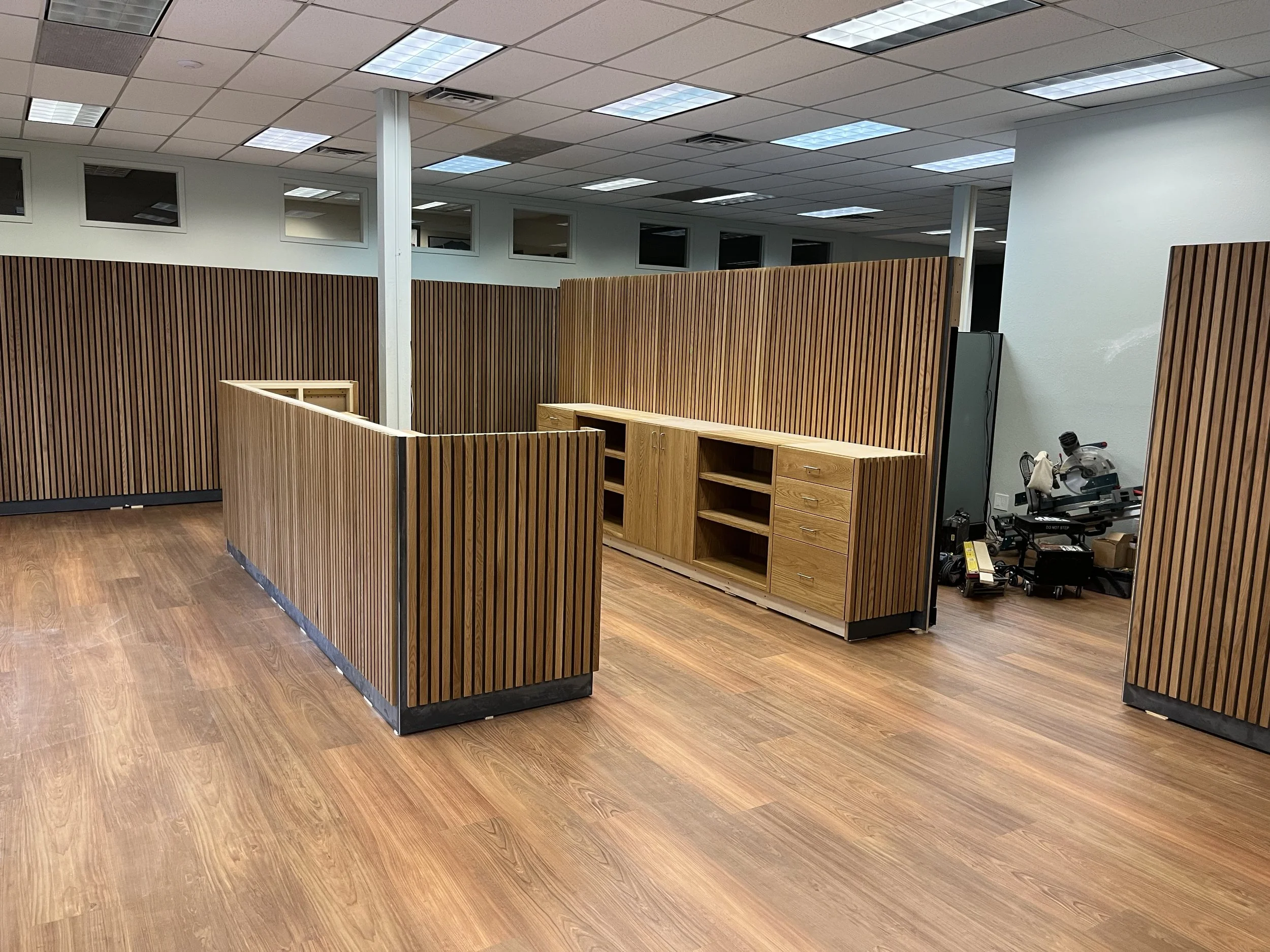 Empty office space with wooden partitions, hardwood floor, ceiling tiles, and fluorescent lighting.