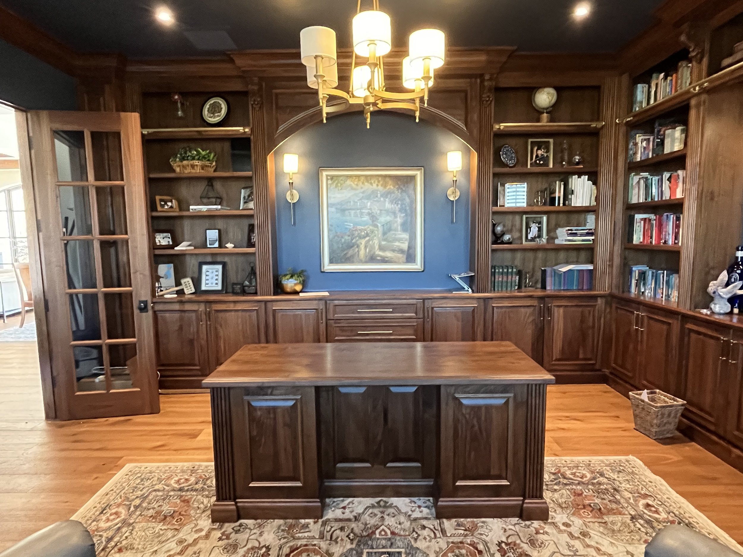 A wooden office or library space with built-in bookshelves, a central desk, and a chandelier hanging from the ceiling. There are various framed pictures and decorative items on the shelves, and a large framed painting on the wall behind the desk.