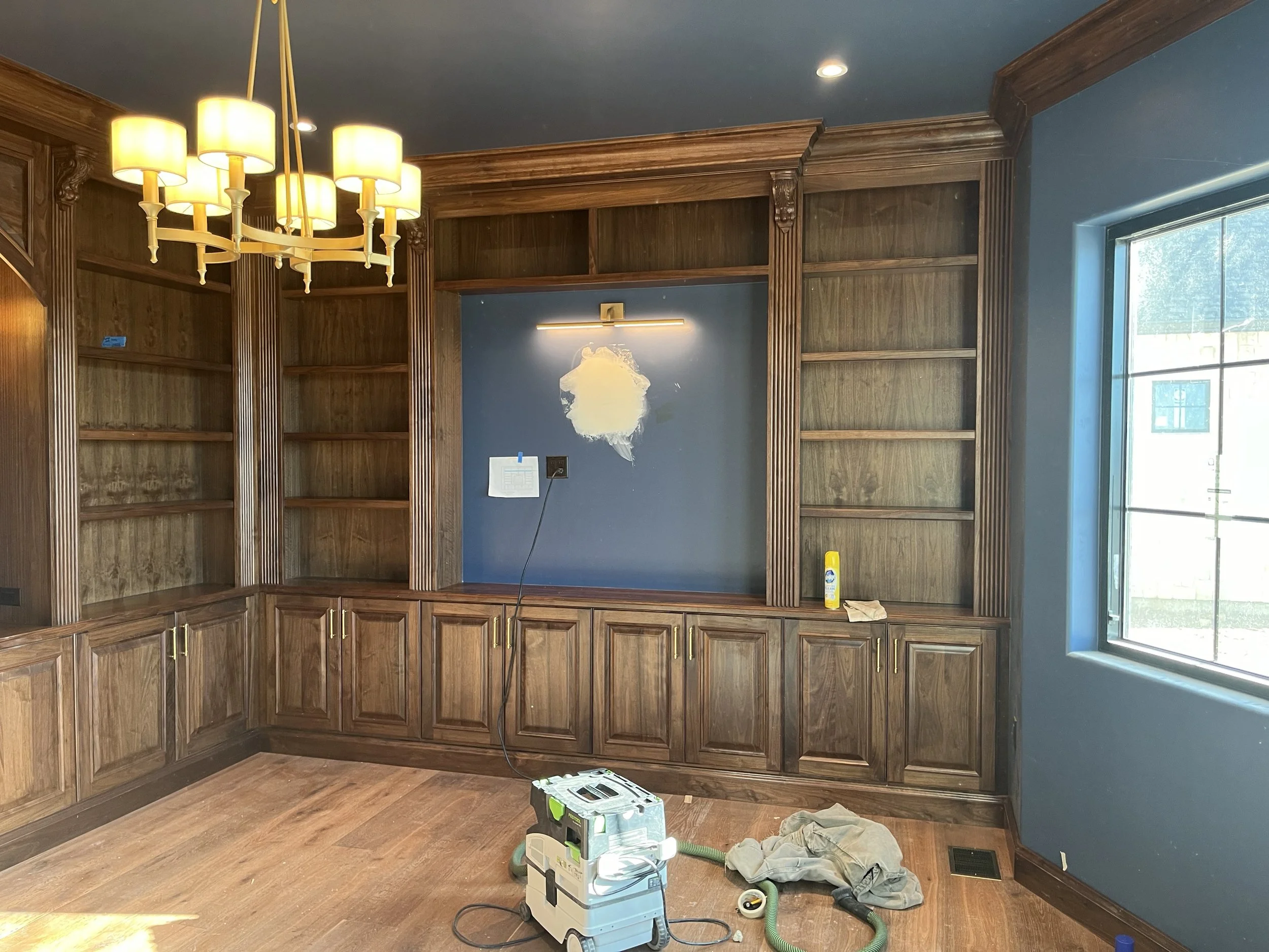 Room with wooden built-in shelves and cabinets, a chandelier, a window, and a blue wall with a light fixture. Cleaning supplies and equipment are on the floor.