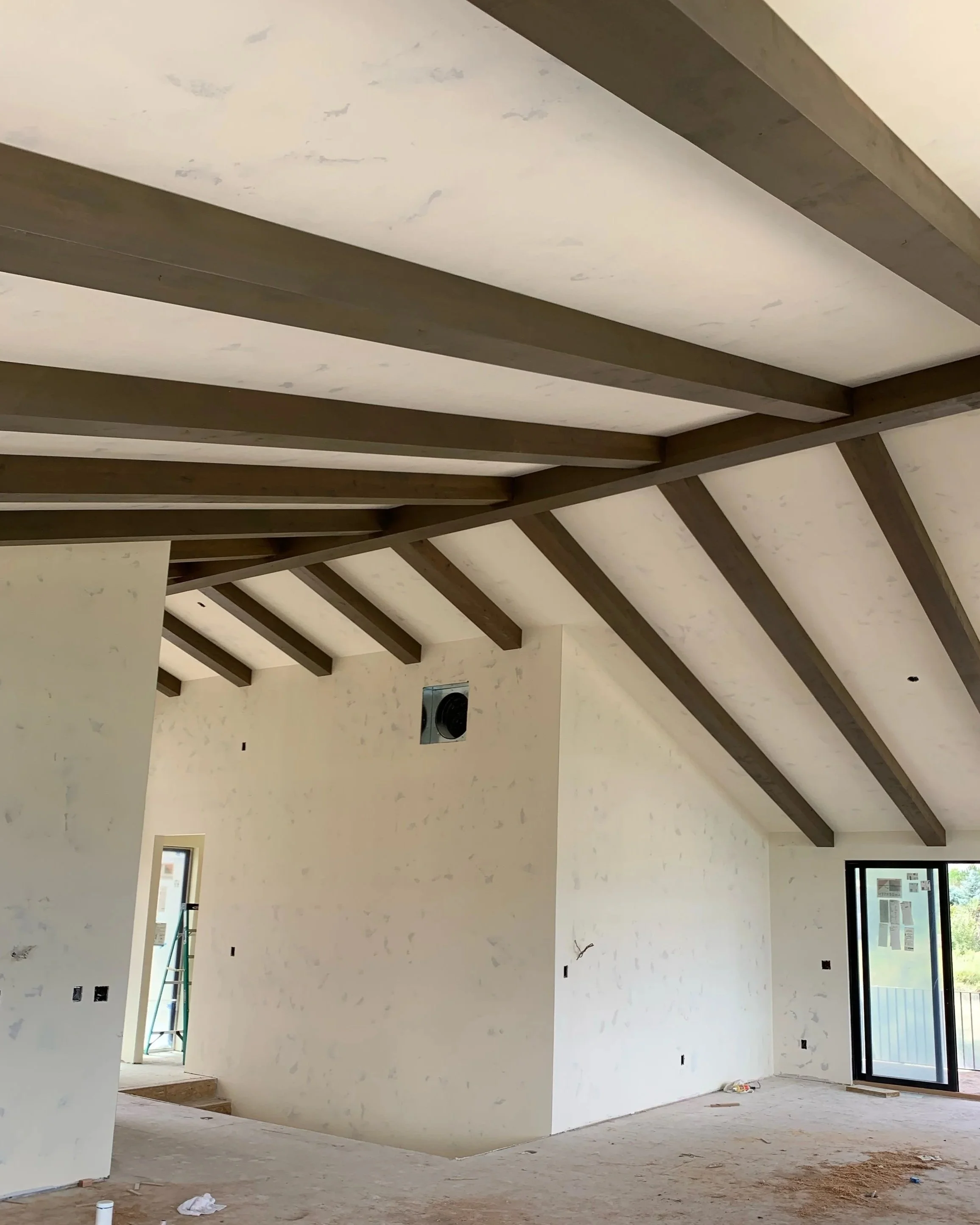Interior of a house under construction with a high ceiling featuring exposed wooden beams and a partially finished wall with windows and a door.