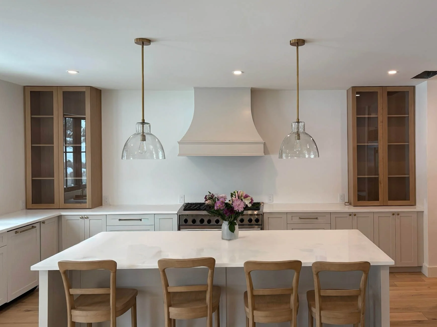 Modern kitchen with white cabinets, a central island with a marble countertop, wooden chairs, a vase of pink flowers, and glass-front cabinets on either side.