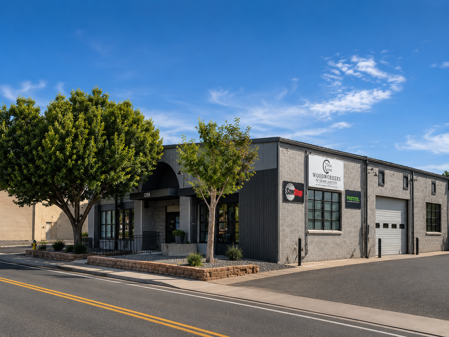 Exterior of a commercial building with signs for Woodworkers of Grand junction CO, SawStop, and Festool, trees, sidewalk, street, and a blue sky with clouds.