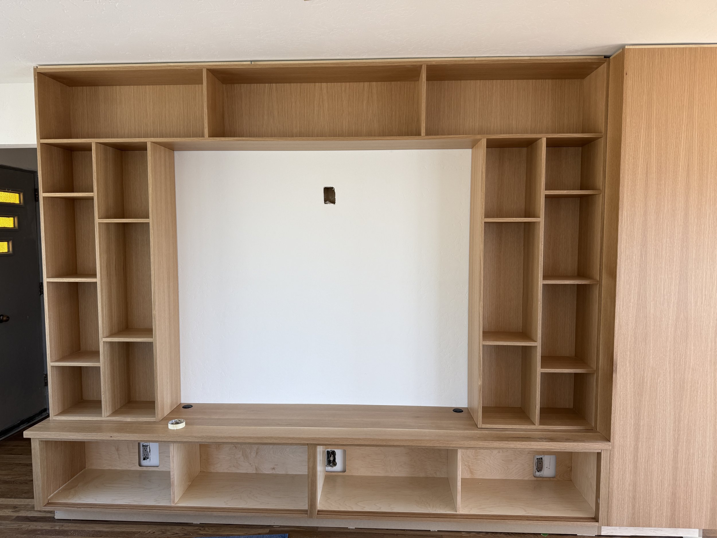 Empty wooden built-in bookshelf with multiple compartments, electrical outlets at the bottom, and holes for wires or fixtures in the wall above.