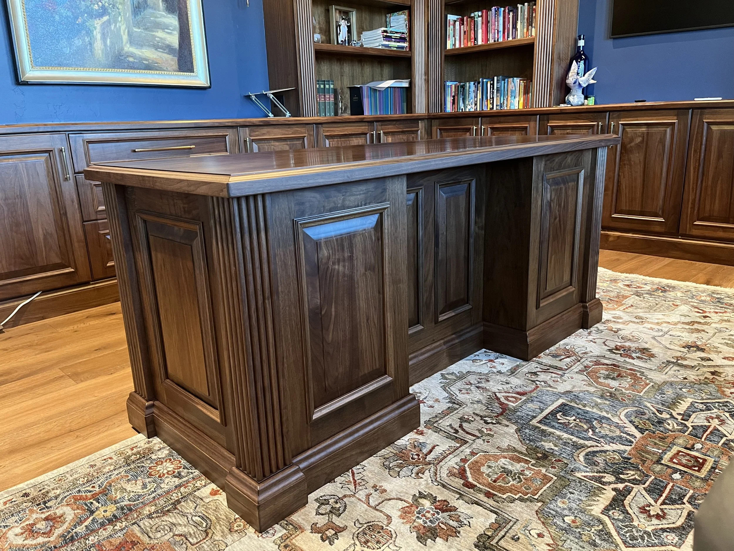 A wooden office desk and bookshelf setup with a colorful area rug on hardwood flooring.