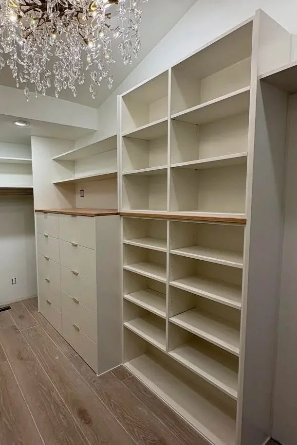 Empty built-in white shelving unit with multiple shelves next to a white cabinet with drawers in a room with wood flooring and a chandelier overhead.