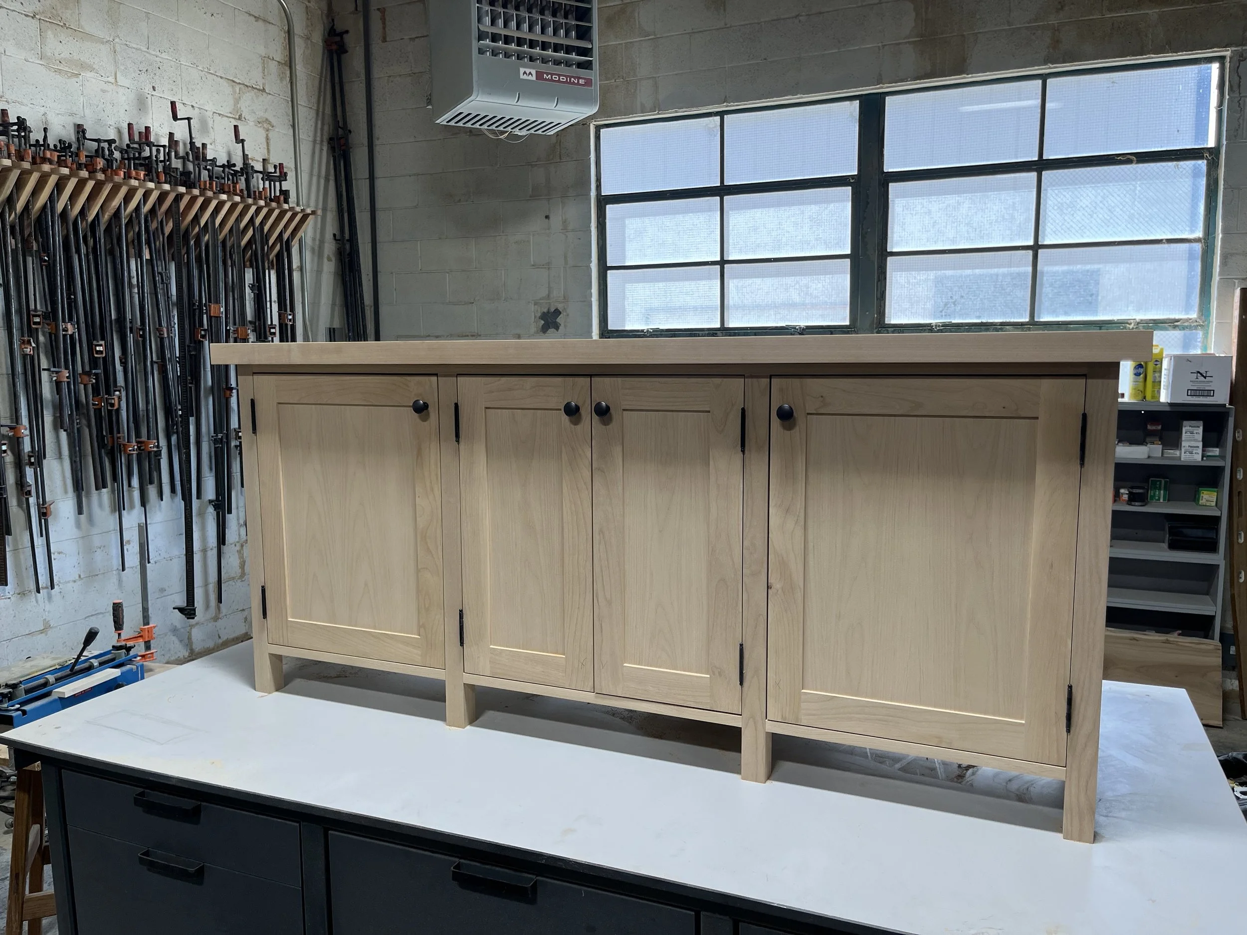 Wooden cabinet with black handles on a white work surface in a woodworking workshop with clamps on the wall and a window in the background.