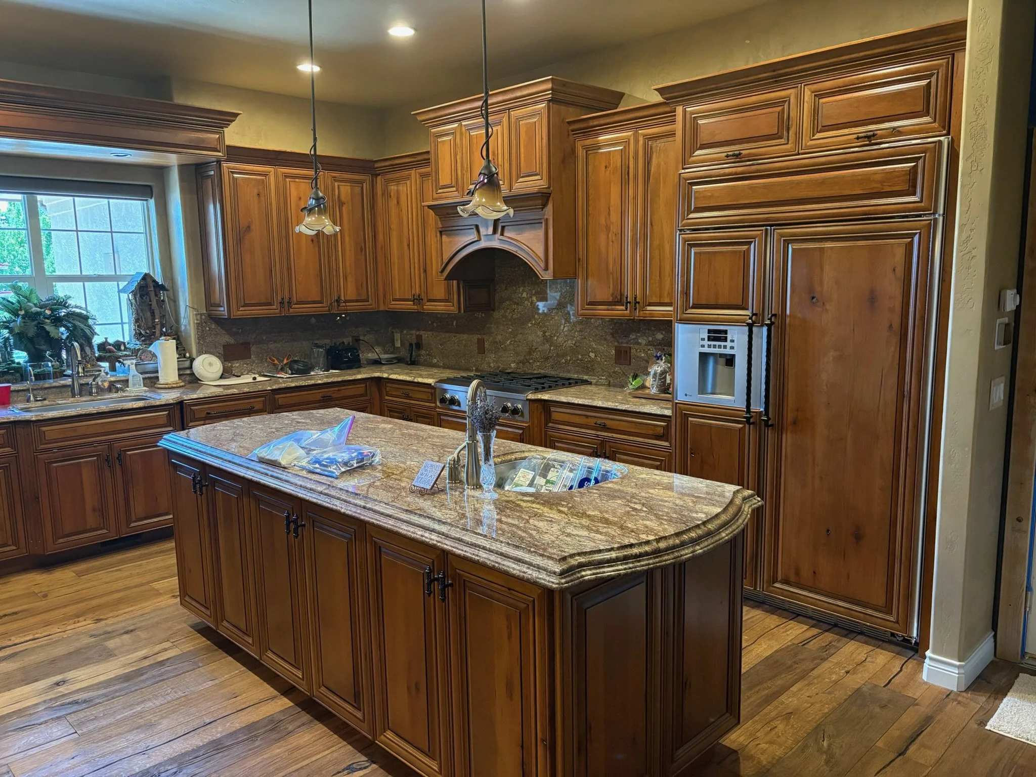 Kitchen with wooden cabinets, granite countertops, an island with a sink, a window above the sink, a refrigerator, and various small appliances and items on the countertops.