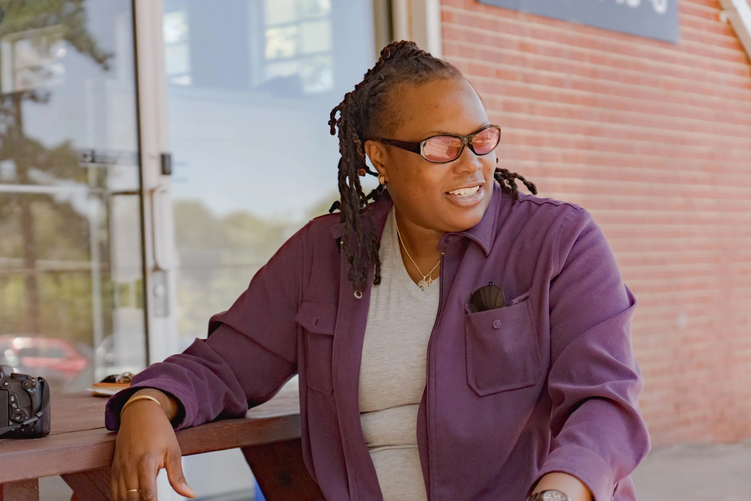 A woman with glasses and dreadlocks sitting at a wooden table outside, smiling and looking to the side.