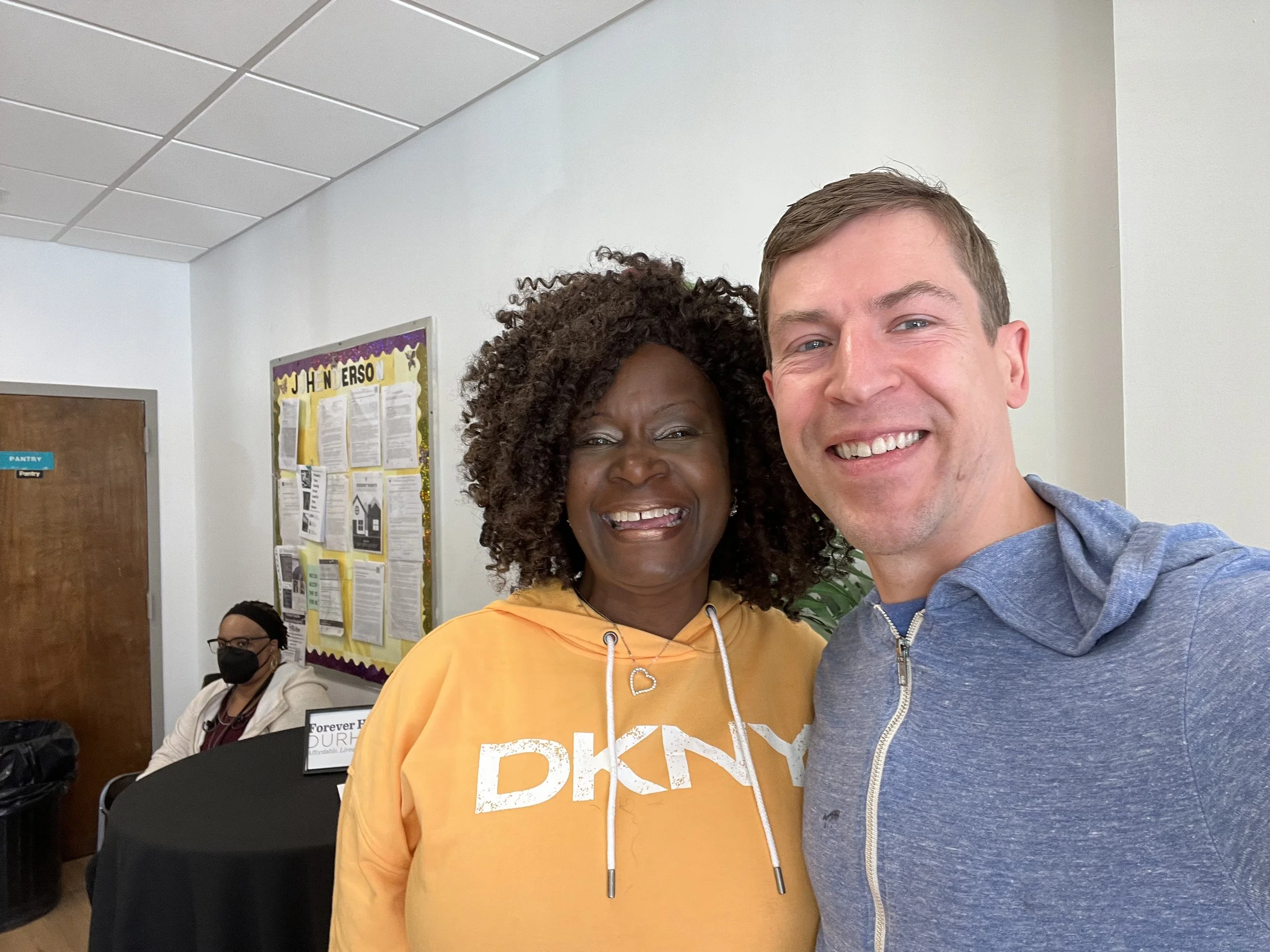 A smiling man and woman taking a selfie inside a room, with a woman sitting and wearing a face mask in the background.