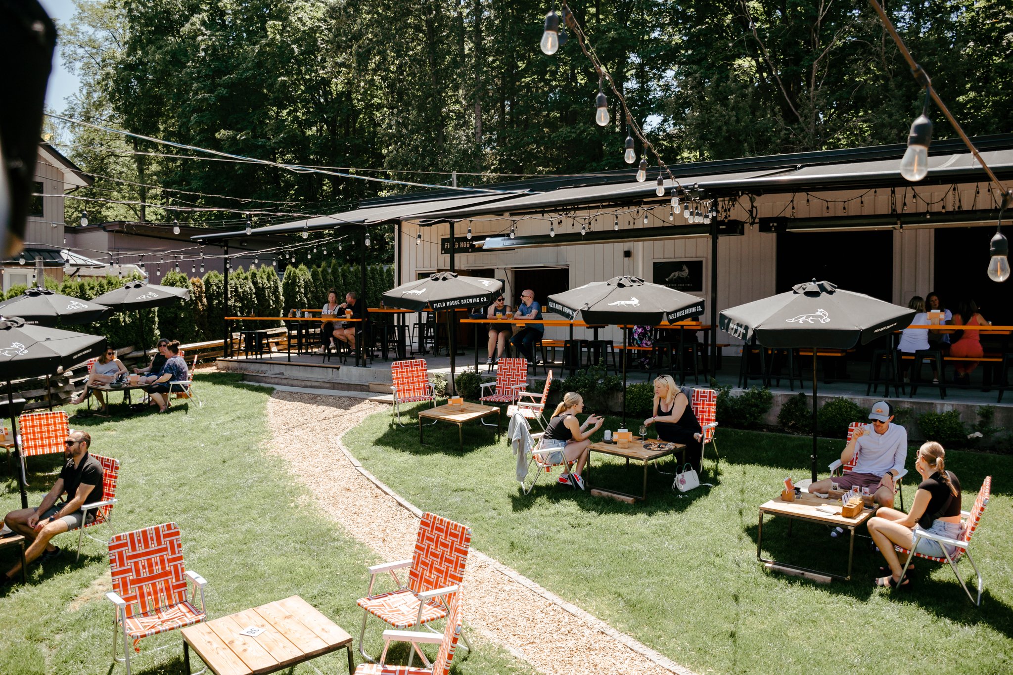 Outdoor patio with people sitting at tables under black umbrellas, surrounded by trees and decorative string lights.