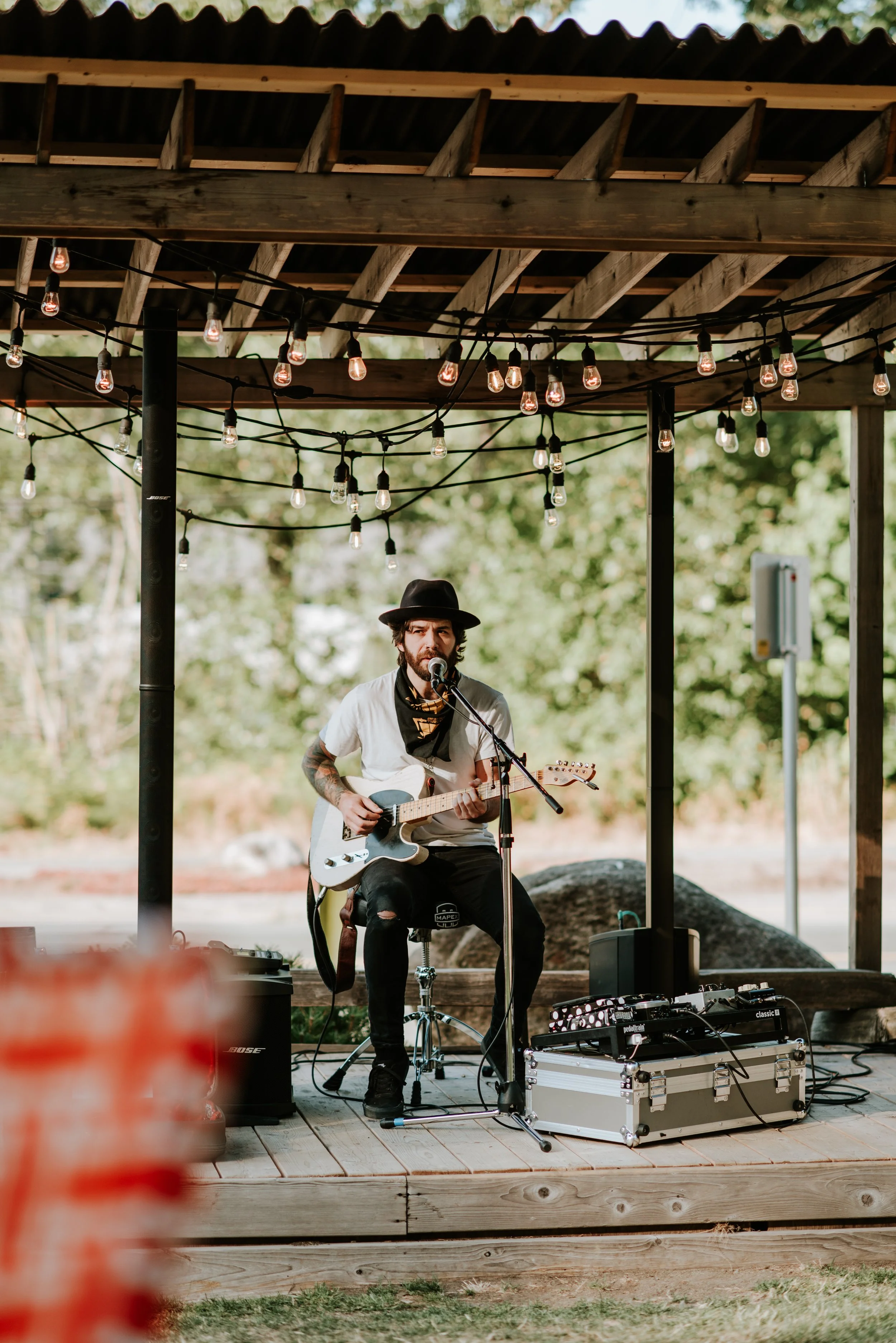 A man with tattoos wearing a black hat and white t-shirt playing an electric guitar and singing into a microphone under string lights on a wooden outdoor stage surrounded by trees.