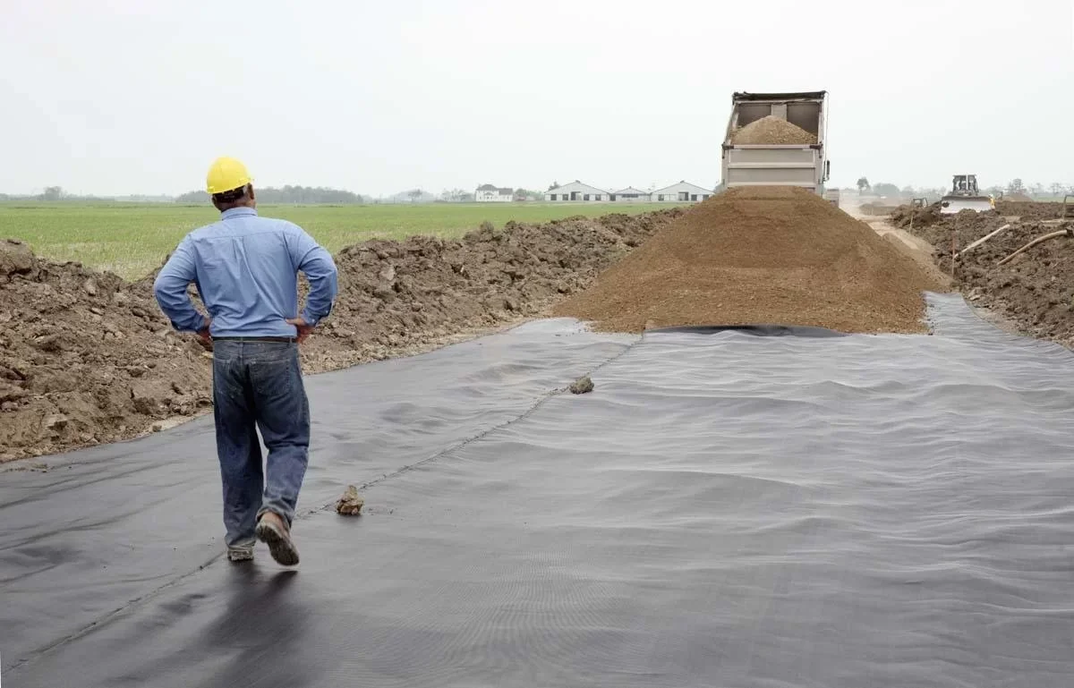 Construction worker wearing a yellow hard hat, blue shirt, and jeans walking on a black Geotextile with a chain, towards a dump truck that is unloading soil in a field.