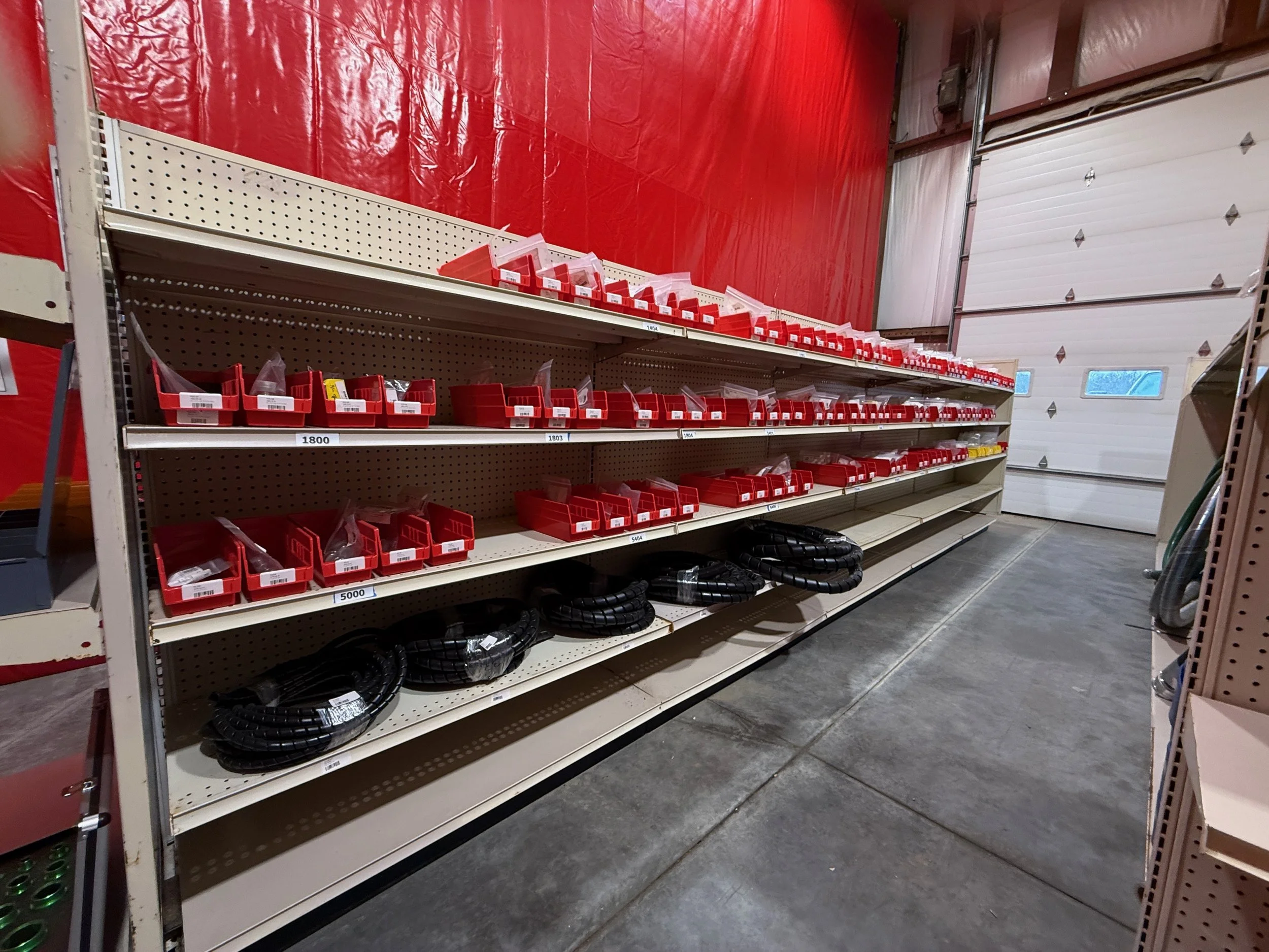 Hydraulic store aisle filled with accessories and hydraulic fittings, store shelves with red bins, some coiled black hoses, and a partially open white garage door, Chester West virginia hydraulic shop aisle