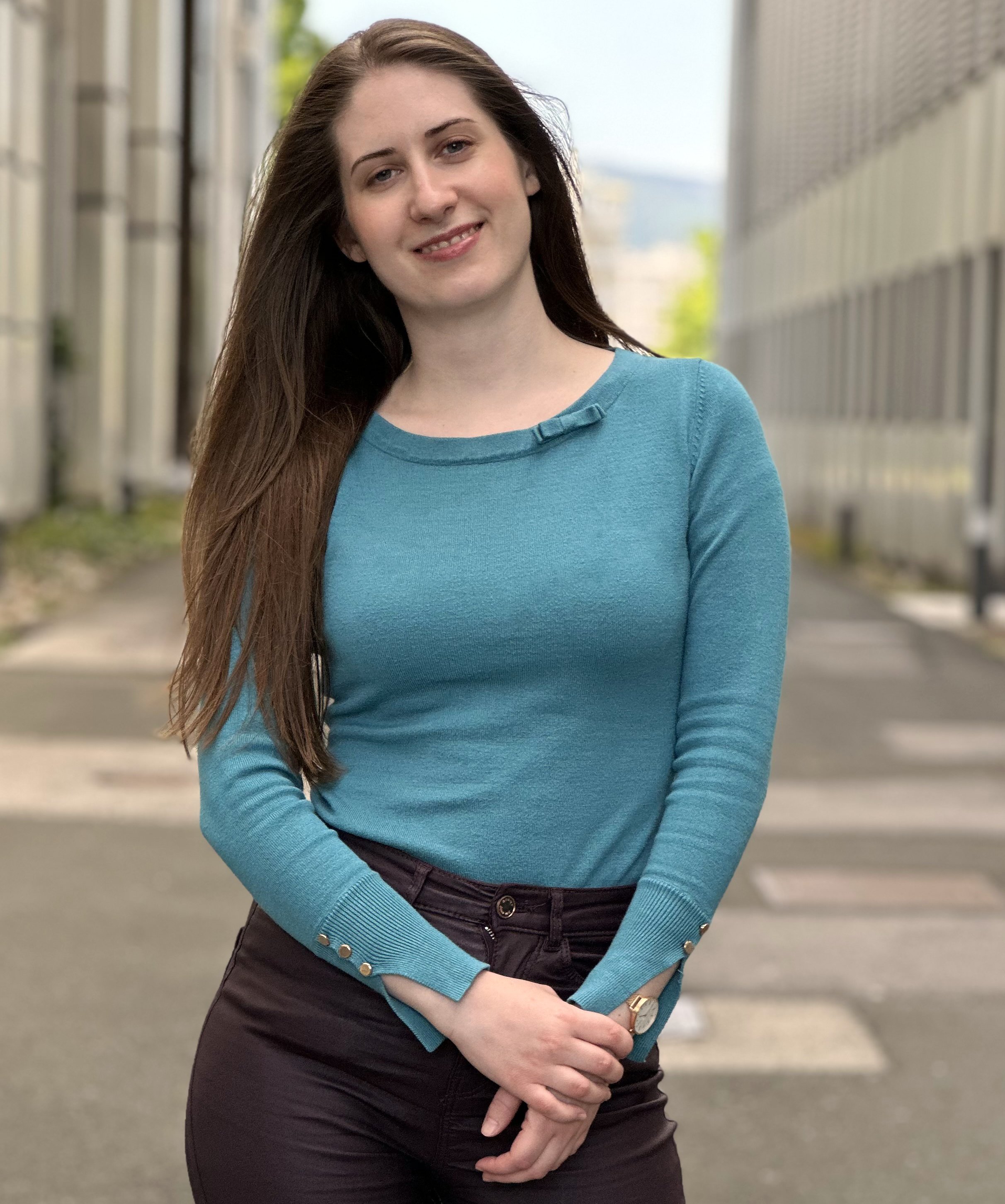 A young woman with long dark hair, wearing a blue long-sleeve top and black pants, standing outdoors with a cityscape background.