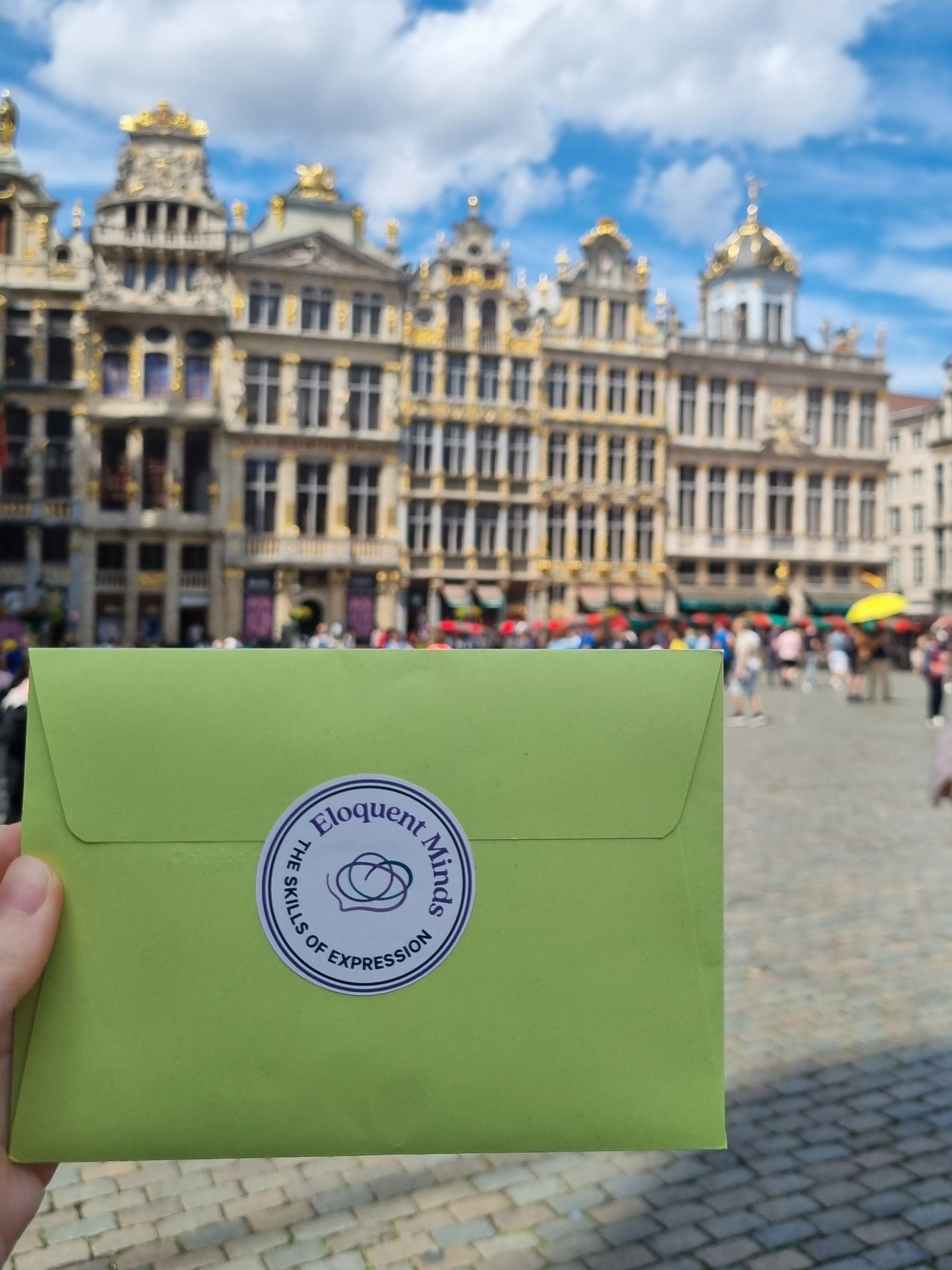 Person holding a green envelope with a logo in front of historic buildings with gold accents in a town square.