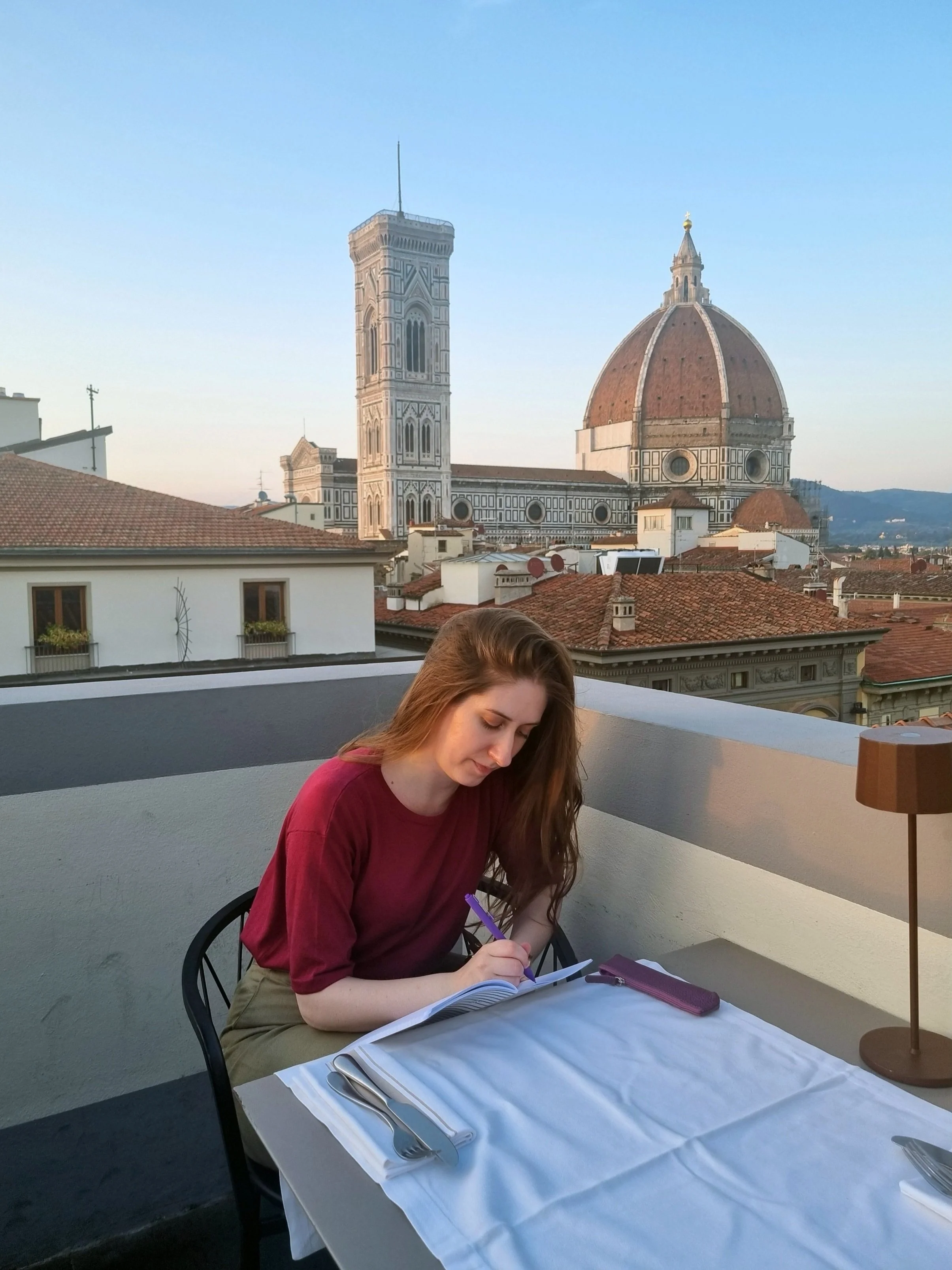 Young woman sitting at an outdoor dining table on a balcony, writing in a notebook with the Florence Cathedral and Giotto's Campanile in the background.