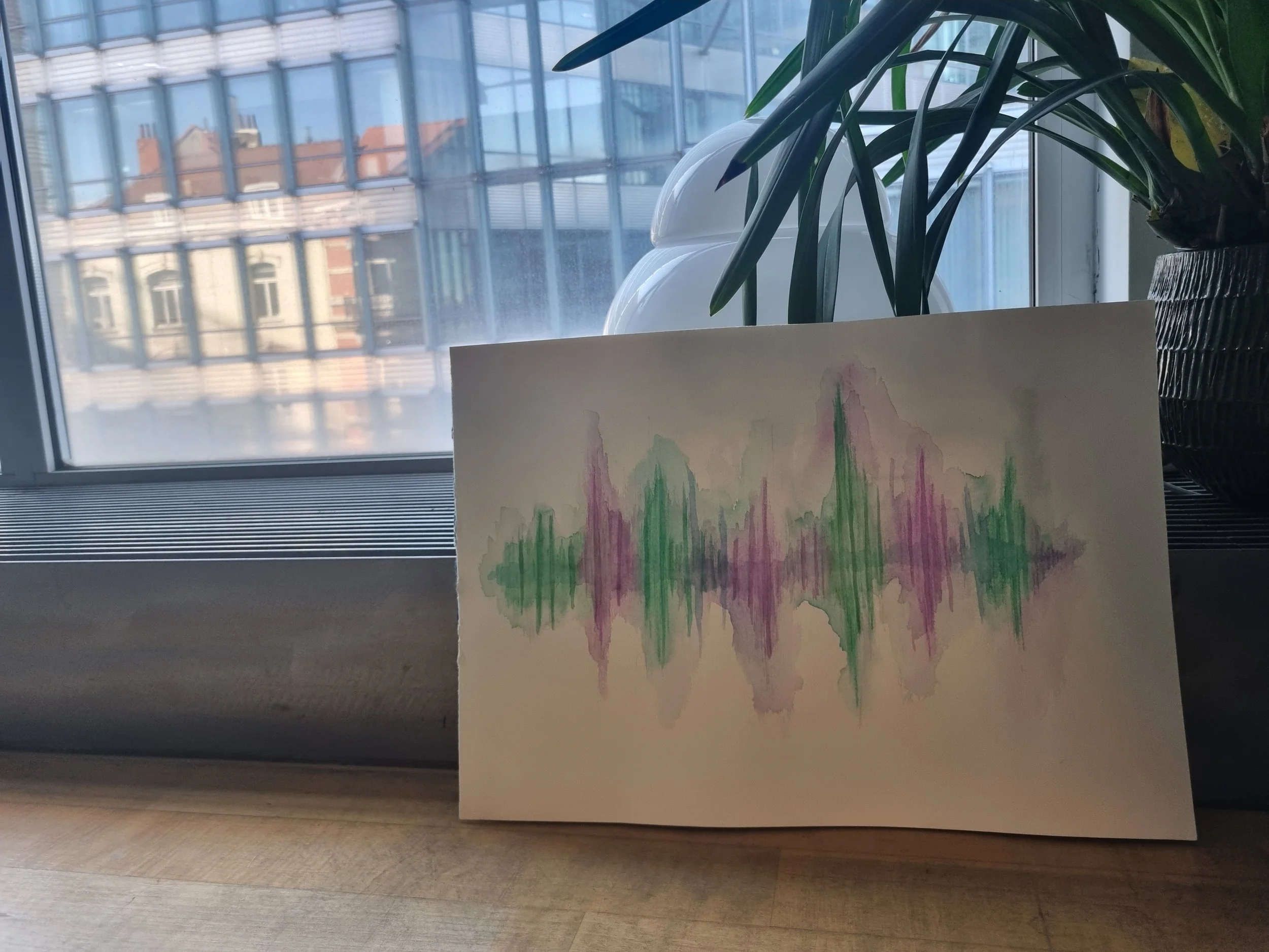 Watercolor painting of colorful sound waves or skyline in green and purple hues, placed on a wooden surface near a window with plants and cityscape outside.