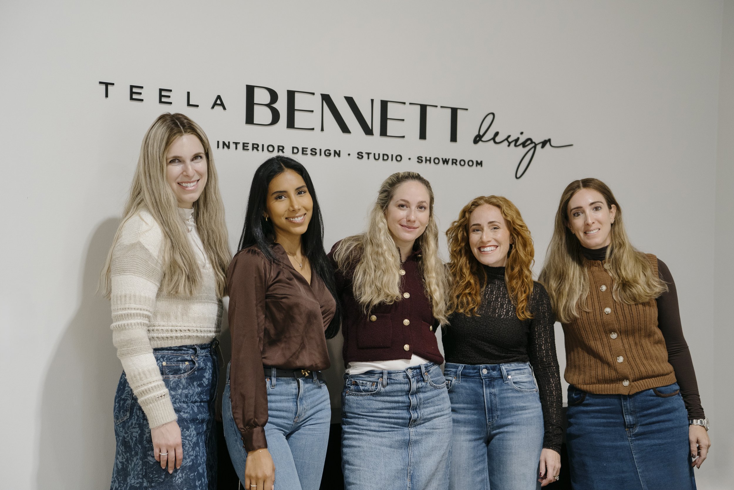 Five women standing in front of a wall with the words 'Teela Bennett Design' and 'Interior Design Studio Showroom.' They are smiling and dressed casually.