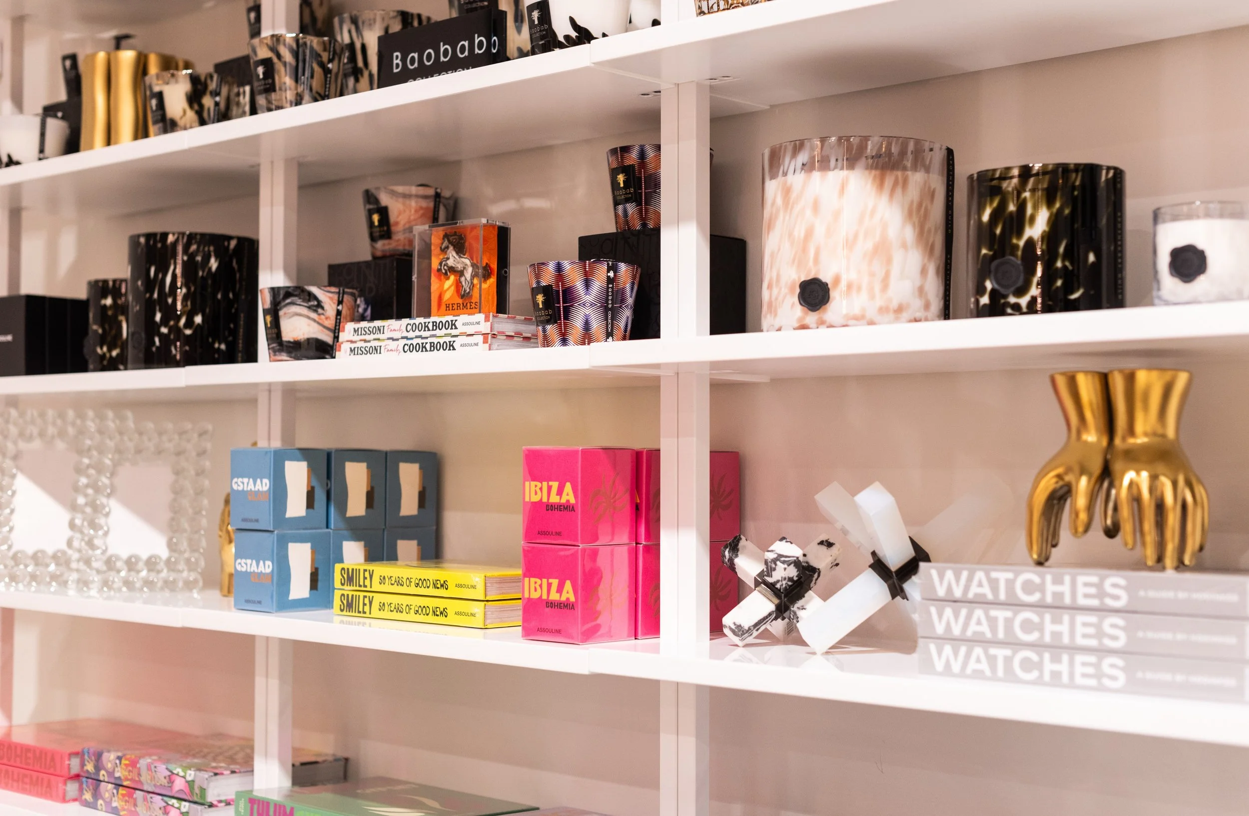 White shelving unit displaying decorative boxes, books, and ornaments including a gold hand sculpture, a white beaded mirror, and a pink box labeled "Ibiza Bohemia."
