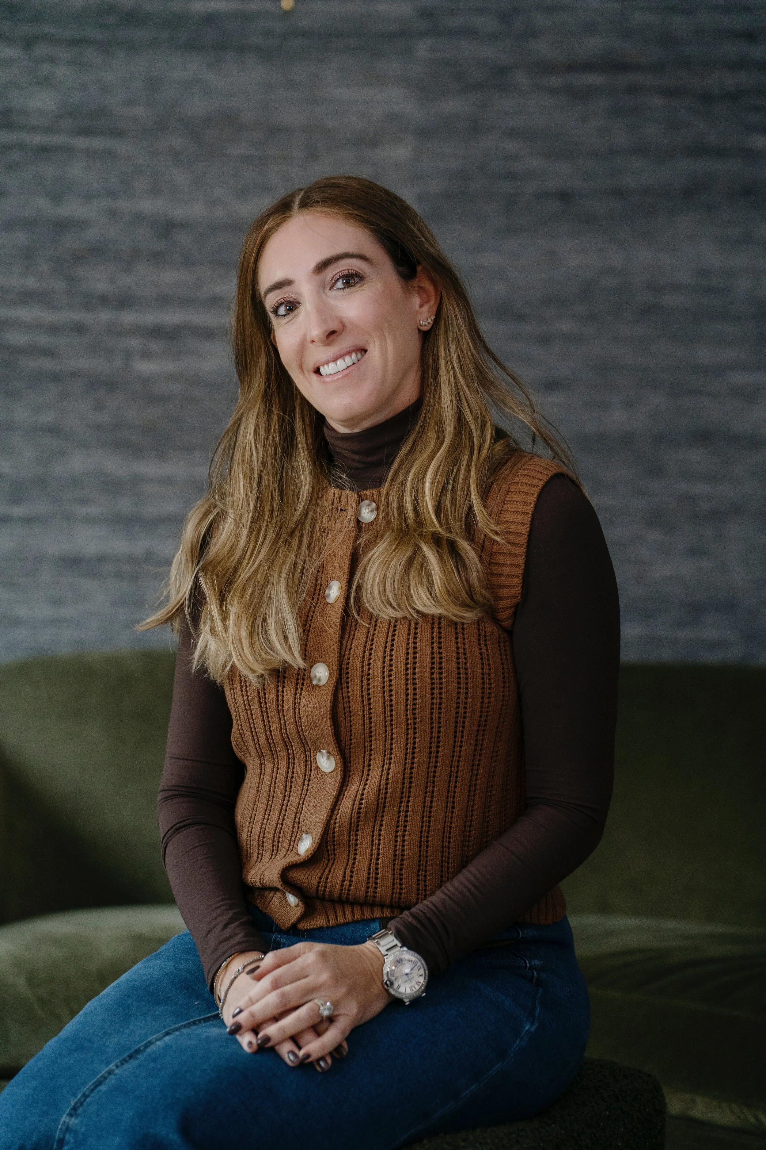 A woman with long, wavy, light brown hair sitting on a green couch, smiling, wearing a brown sleeveless knit vest with pearl buttons over a dark turtleneck, blue jeans, a watch, and rings.