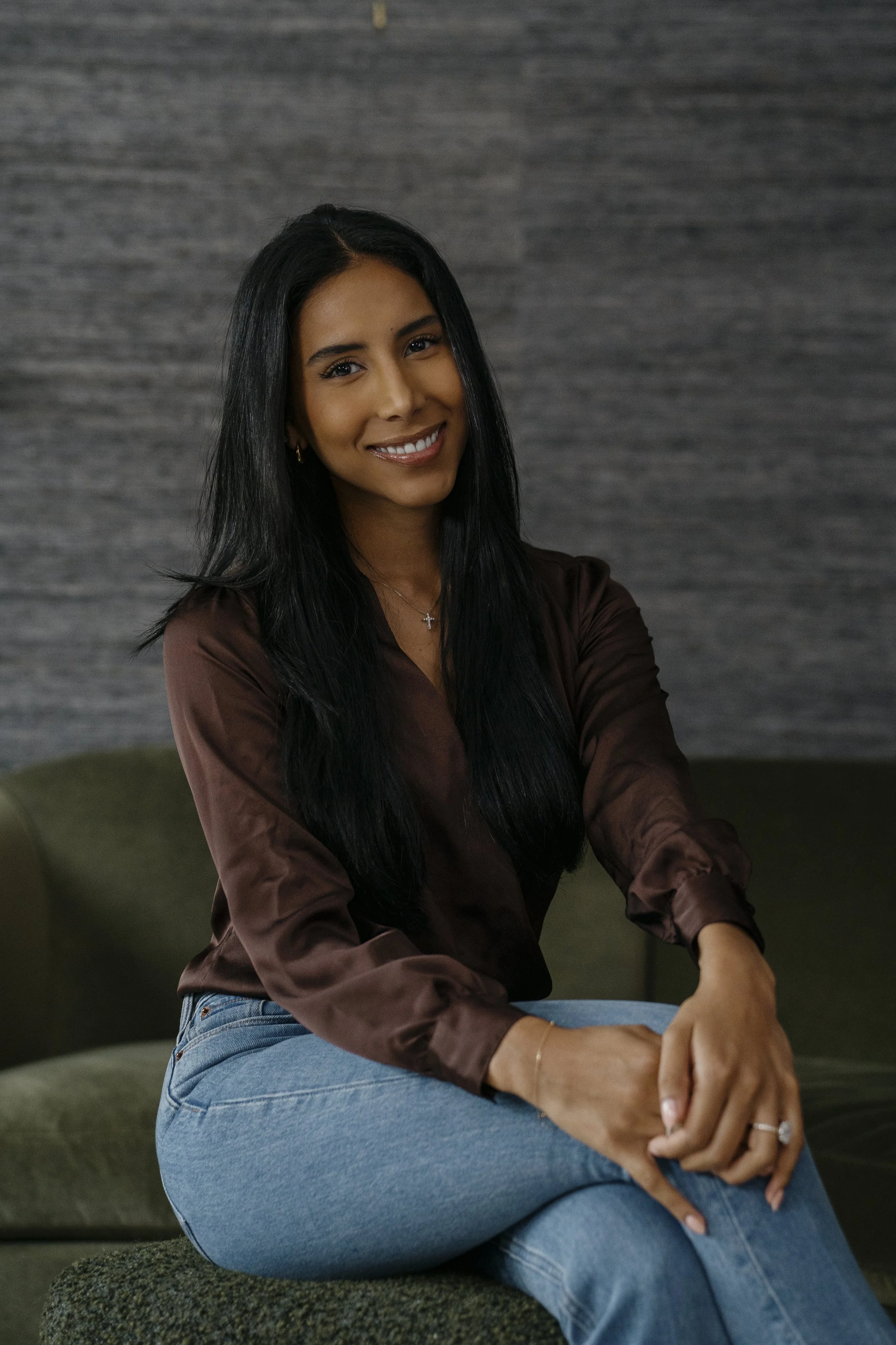 A woman with long black hair, smiling, wearing a brown silk blouse and light blue jeans, sitting on a green sofa against a textured gray wall.