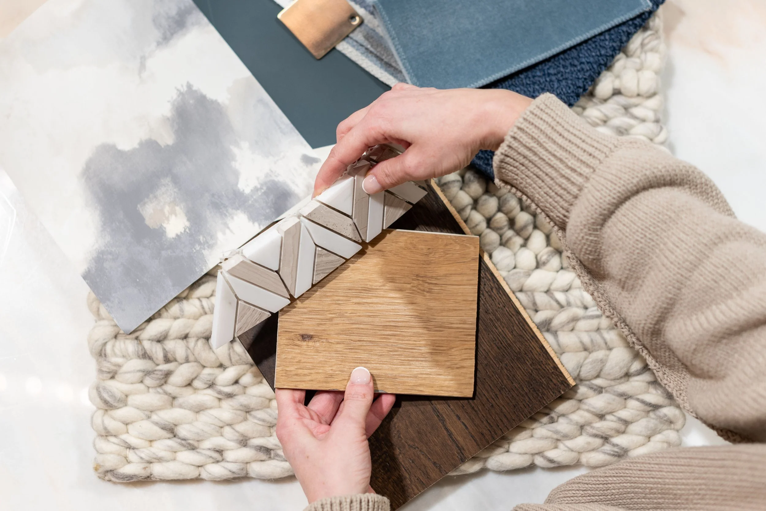 A person is arranging decorative tiles on a wooden sample board, surrounded by fabric samples, with a textured white blanket underneath.
