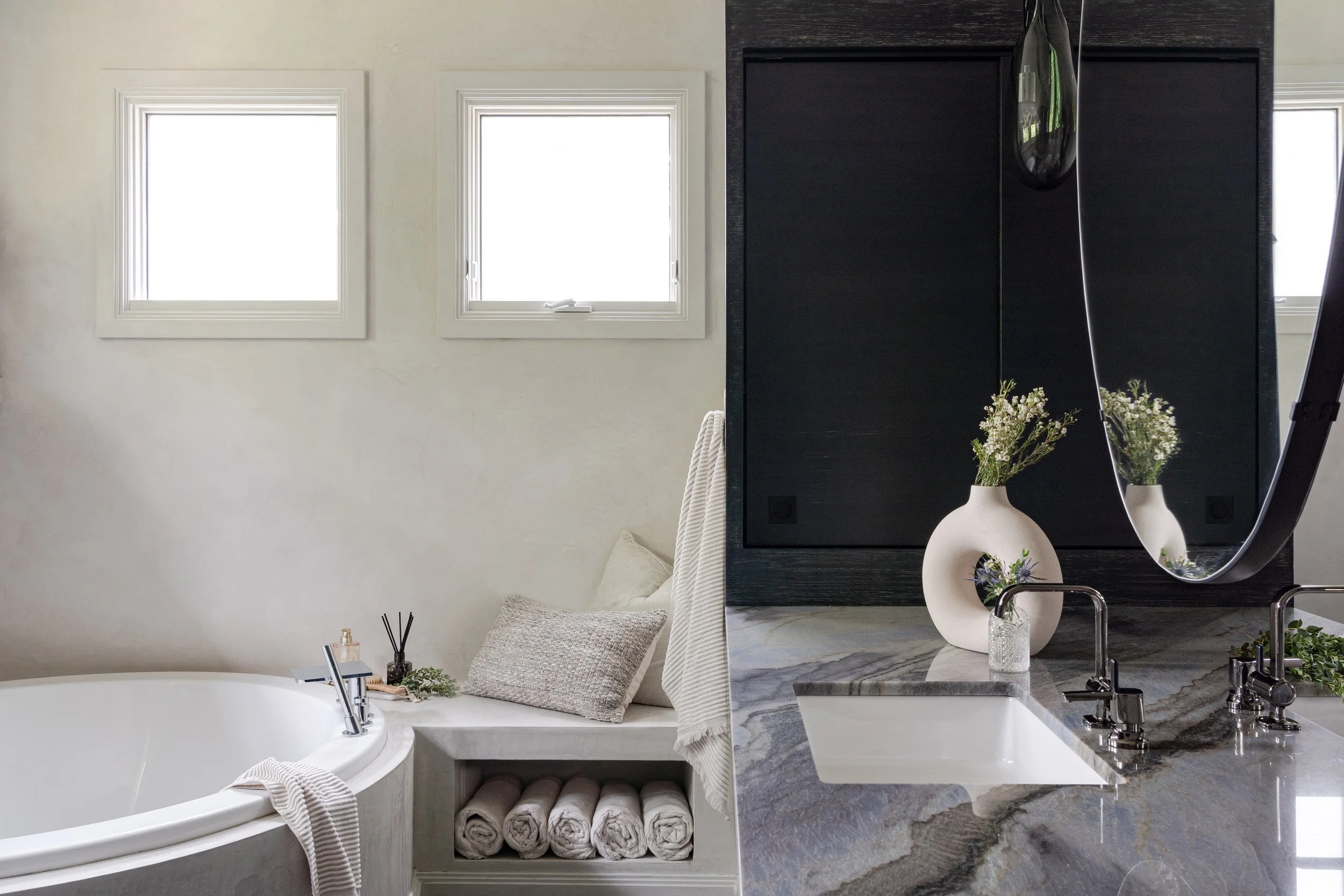 Split view of a bathroom. Left side has a white bathtub with a striped towel and a pillow, next to a small table with a bottle, reed diffuser, and greenery. Right side has a marble countertop with a sink, a white vase with flowers, a soap dispenser, and a mirror with a hanging pendant light above.