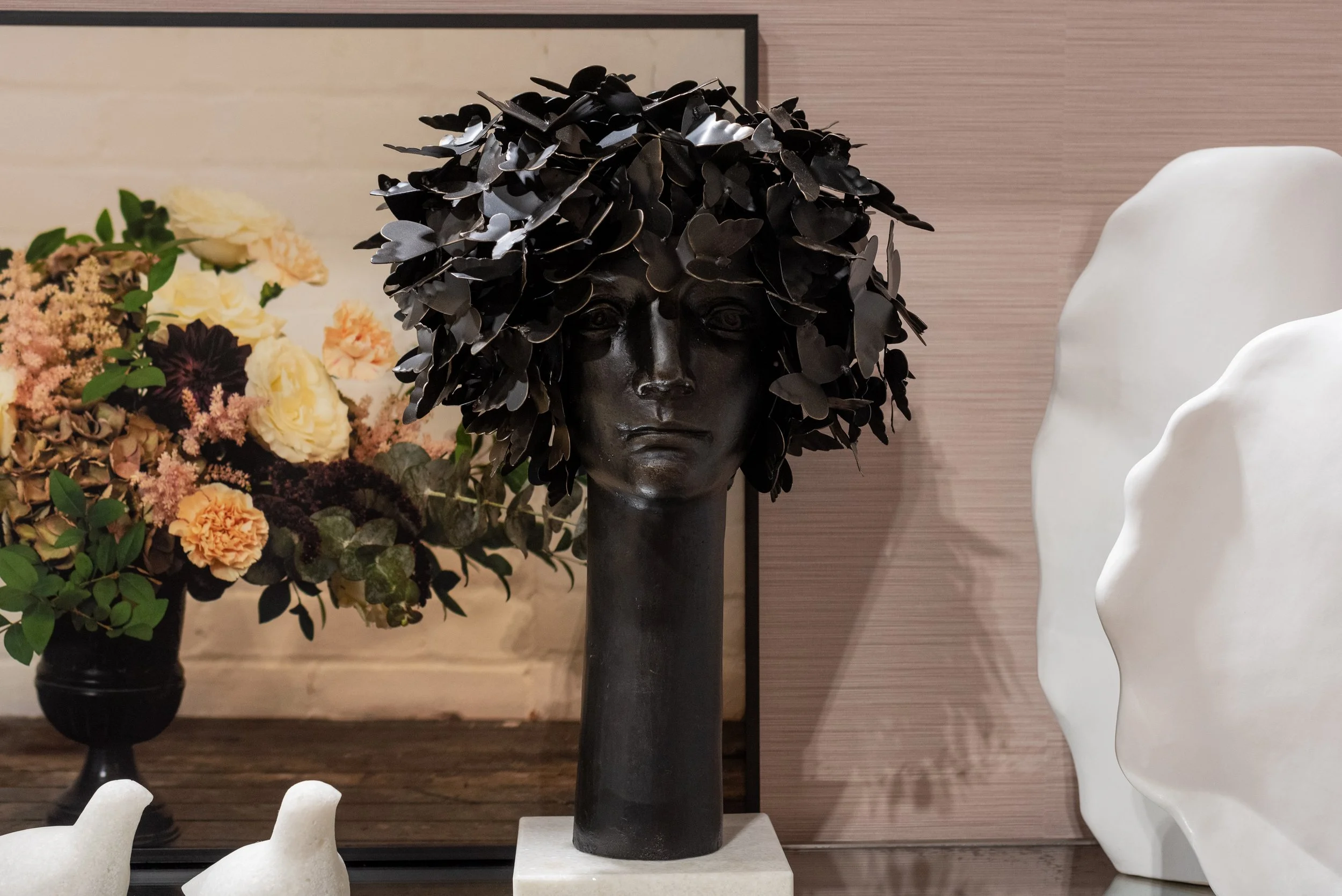 Decorative black bust sculpture of a woman's head with leaves as hair, displayed on a white pedestal. Background includes a floral arrangement with various flowers and leaves, and some white abstract sculptures to the right.
