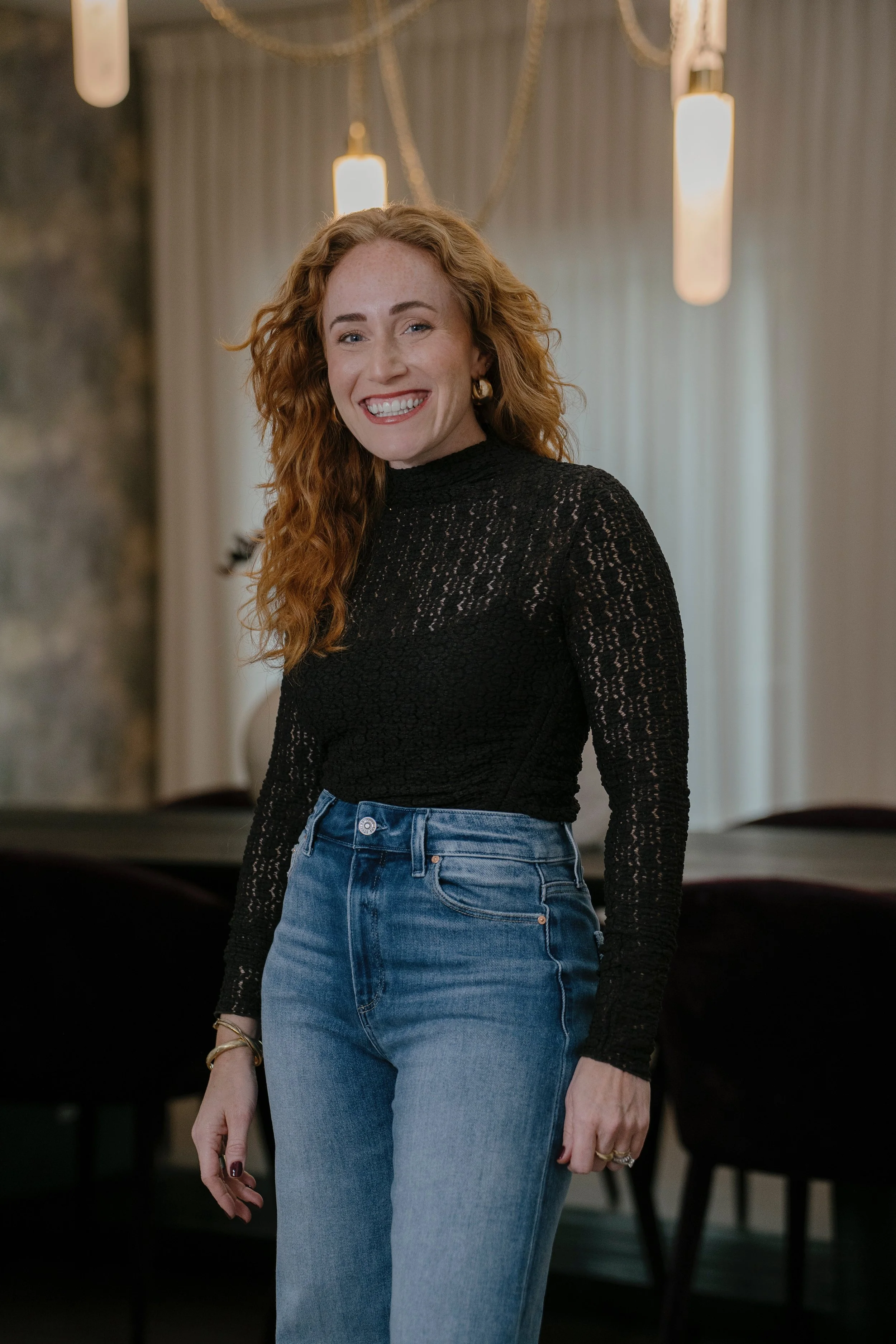 A woman with curly red hair smiling, wearing a black lace long-sleeve top and high-waisted blue jeans, standing in a modern interior space with pendant lighting and curtains in the background.
