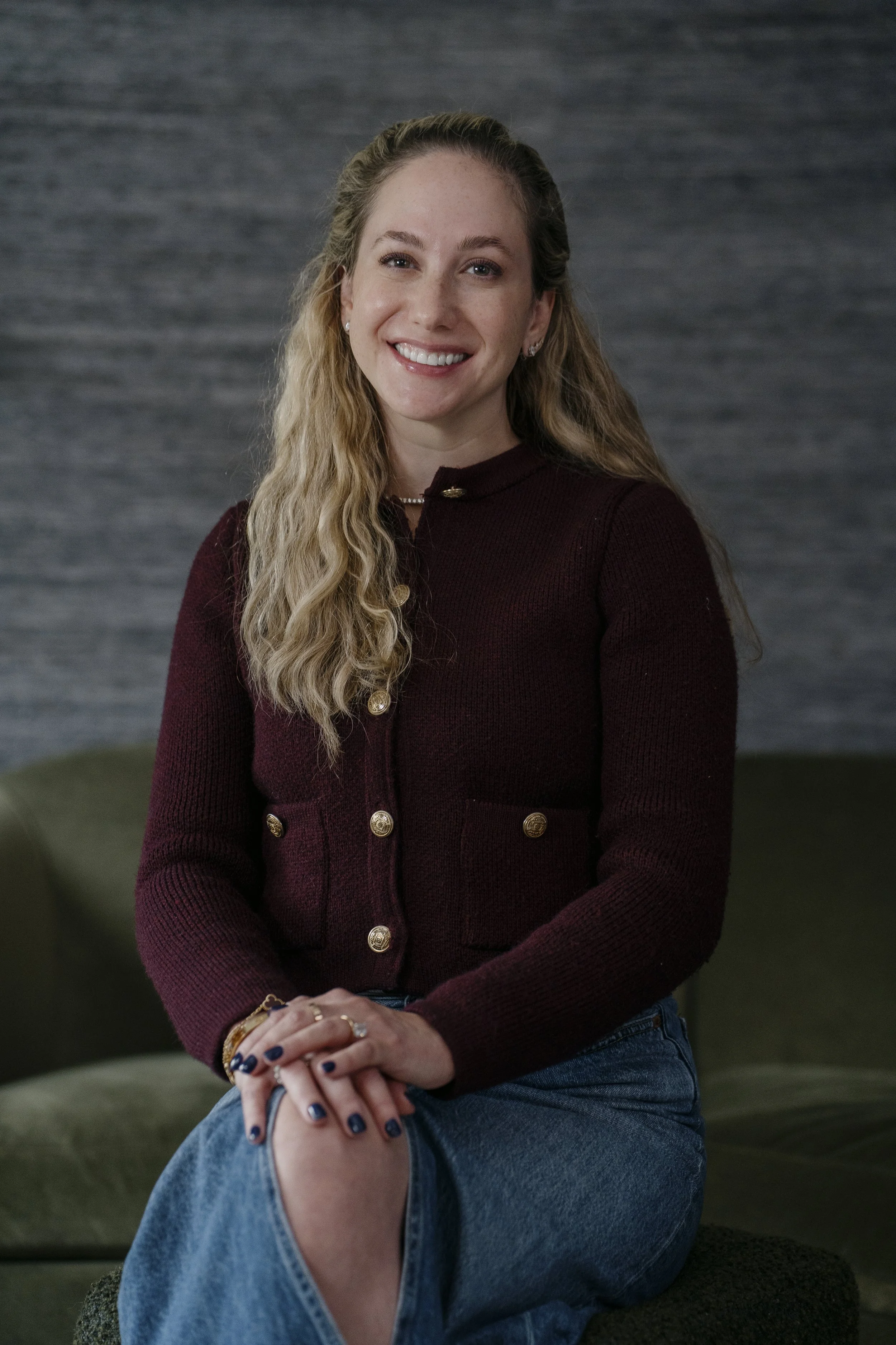 A young woman with long blonde hair and blue eyes, smiling, sitting on a dark green couch against a grey textured wall, wearing a burgundy jacket with gold buttons and blue jeans.
