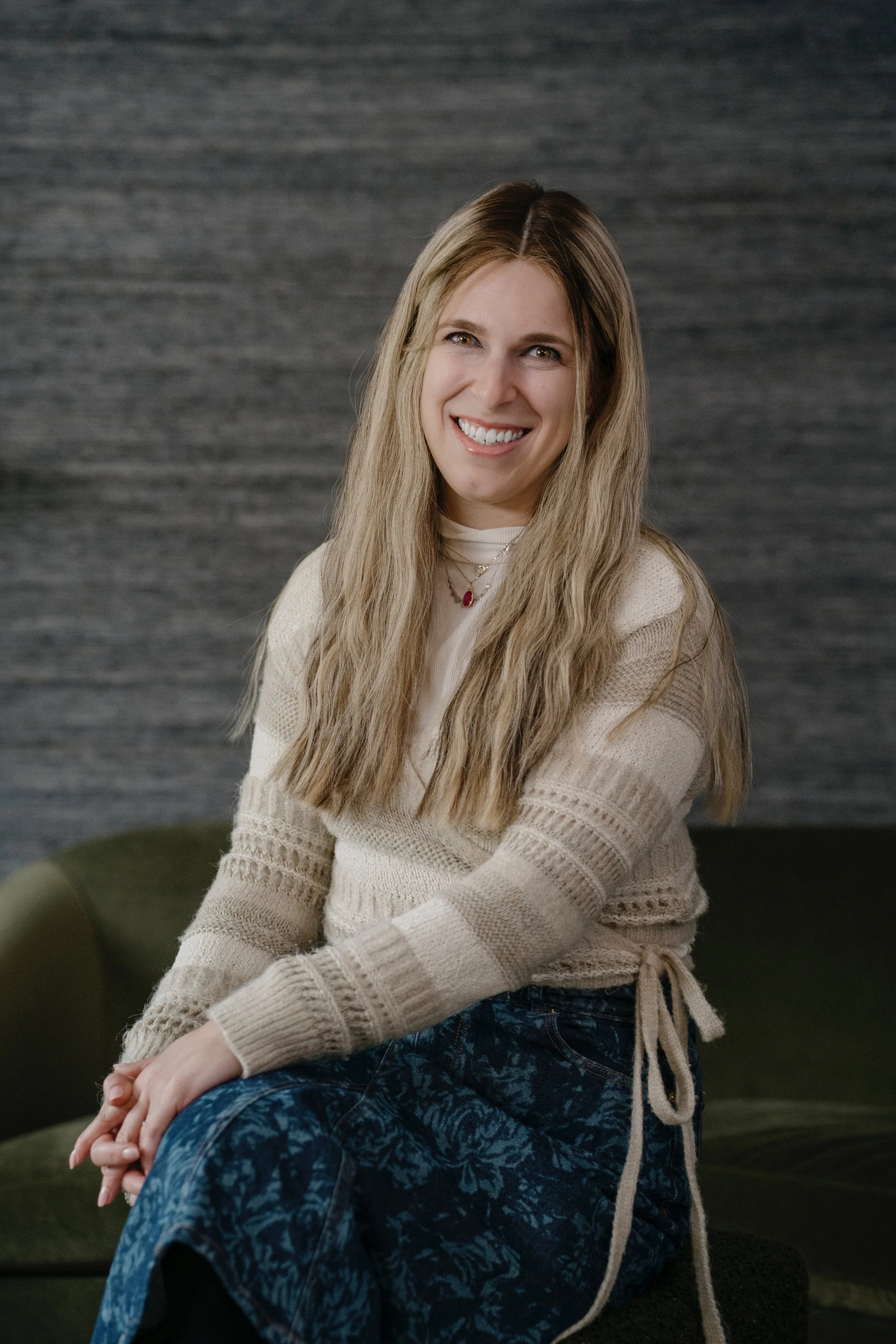 A woman with long blonde hair smiling and sitting on a green couch against a gray textured background.