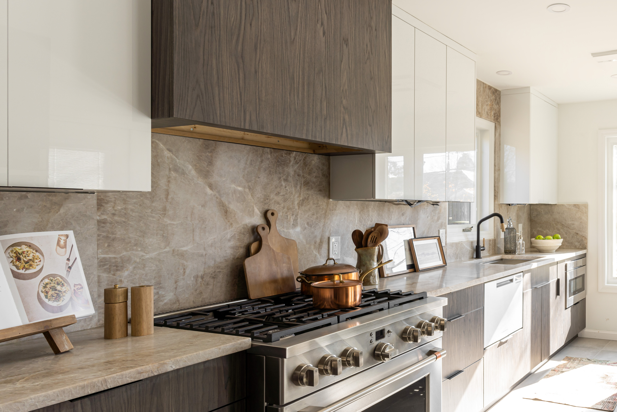 Modern kitchen with beige marble countertops, dark wood and white cabinetry, copper pots on stove, framed photos and kitchen utensils, window with natural light, and green apples in a white bowl.